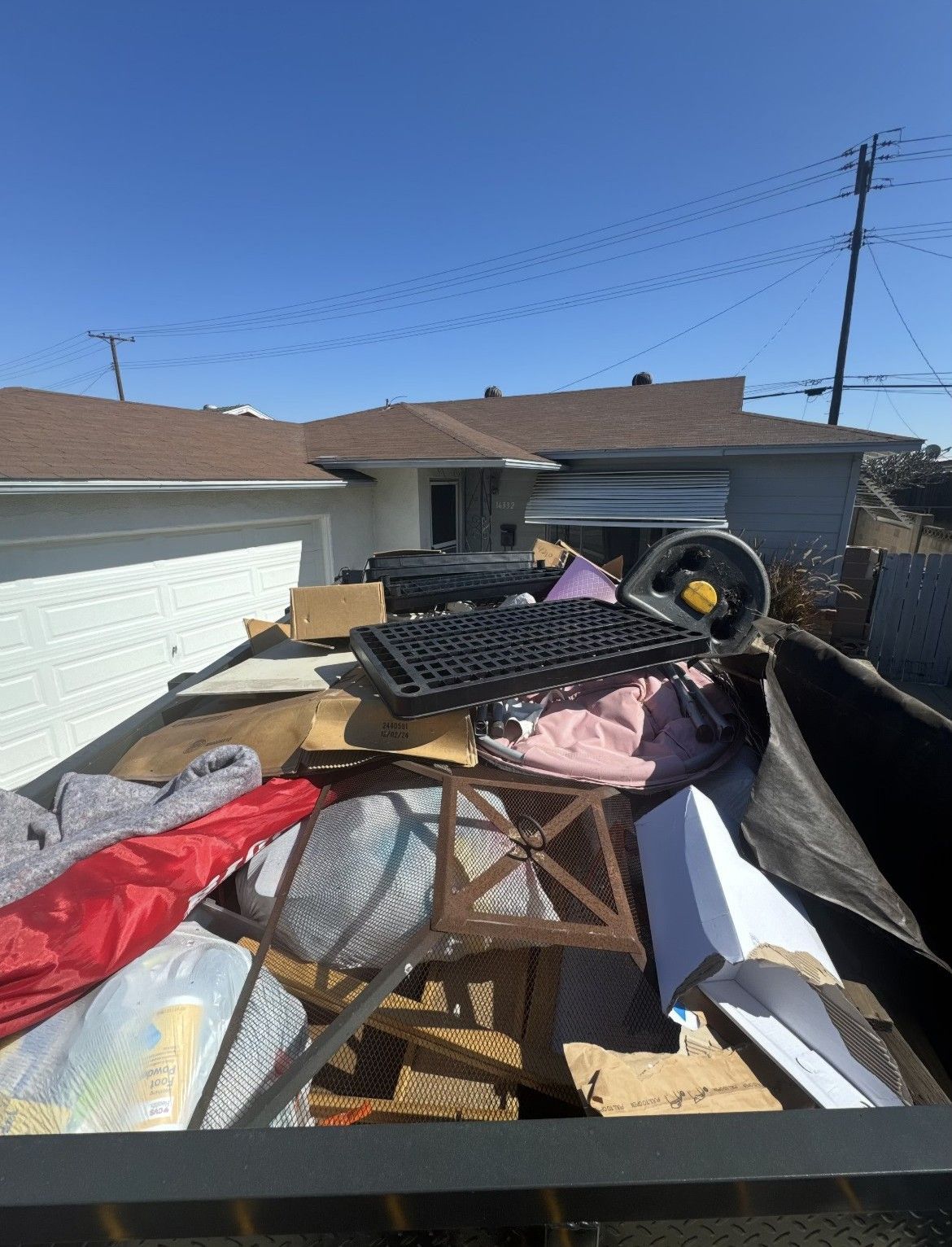 A dumpster overflowing with various trash items, including furniture, cardboard, and blankets; a sunny outdoor setting.