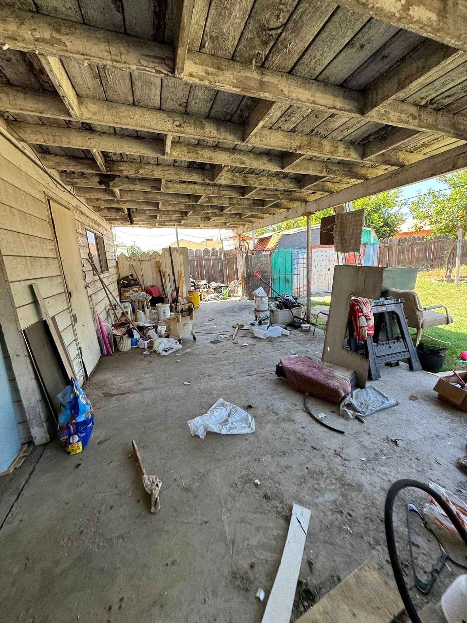 Cluttered outdoor area under a weathered  roof. Debris, discarded items, and a bicycle are scattered on the concrete floor.