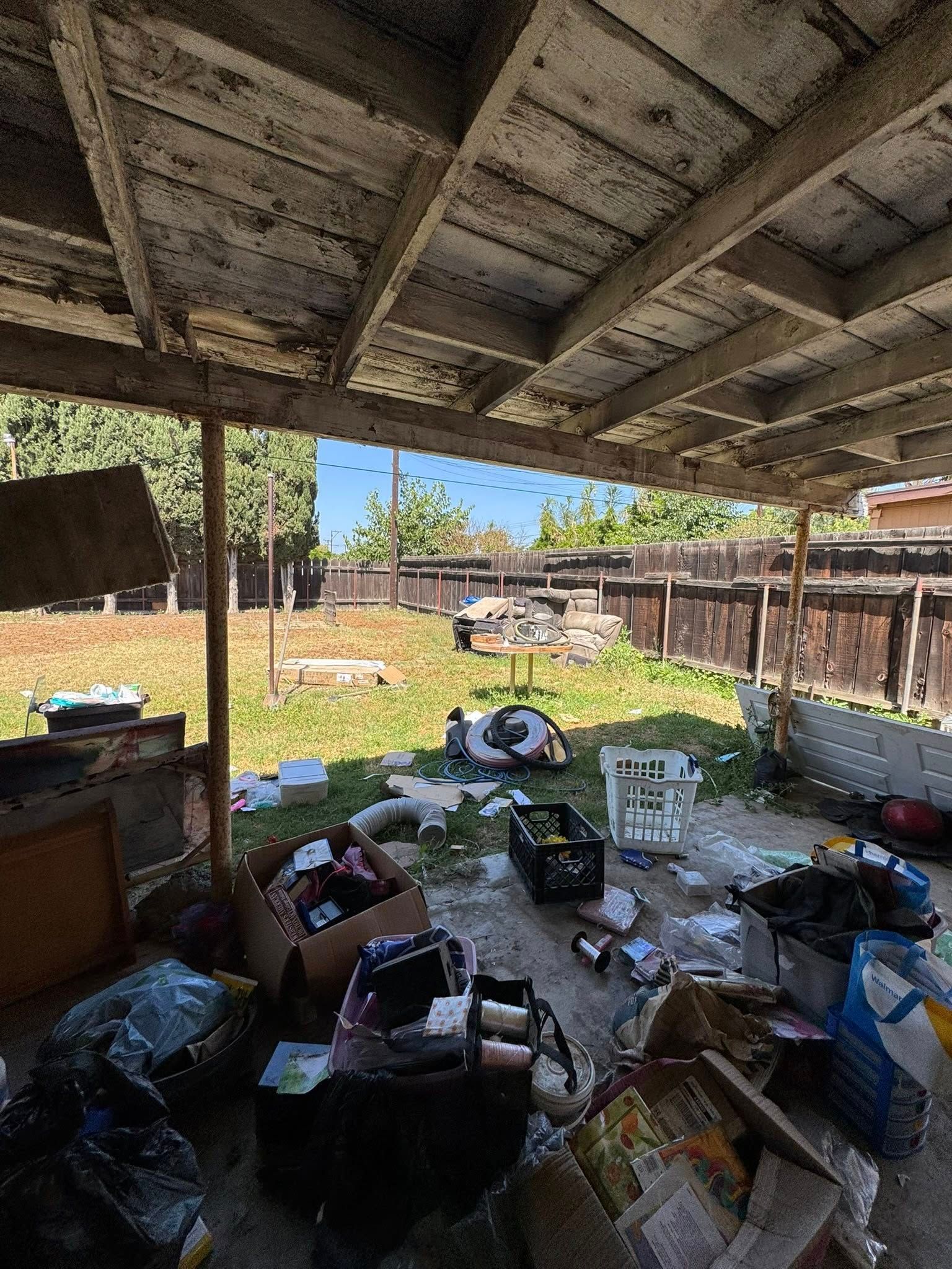 Messy backyard under a deteriorating roof. Trash, debris cover the ground; a wooden fence in the background.