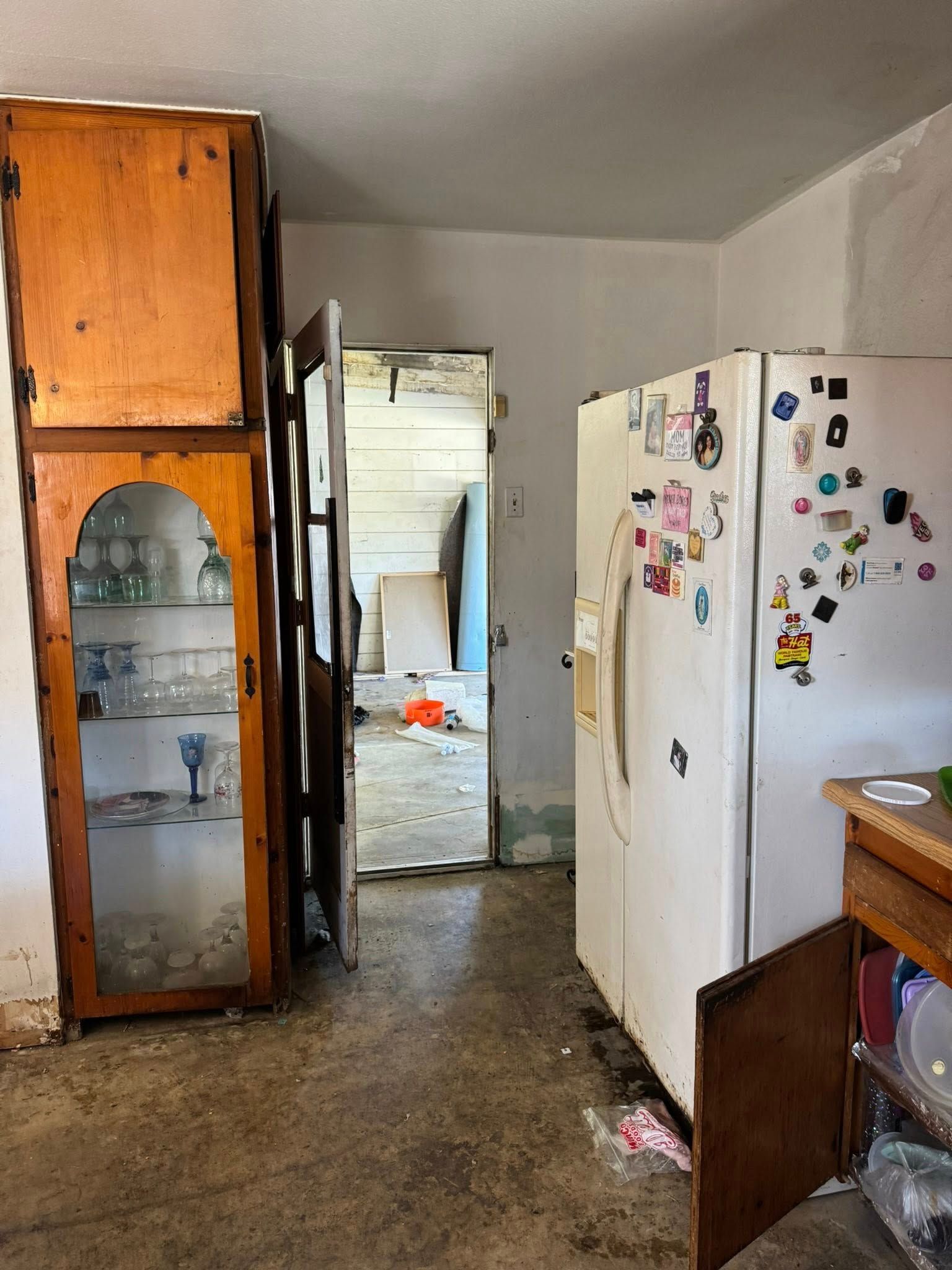 Kitchen with a refrigerator, cabinet, and an open doorway to another room. Dirty floor.
