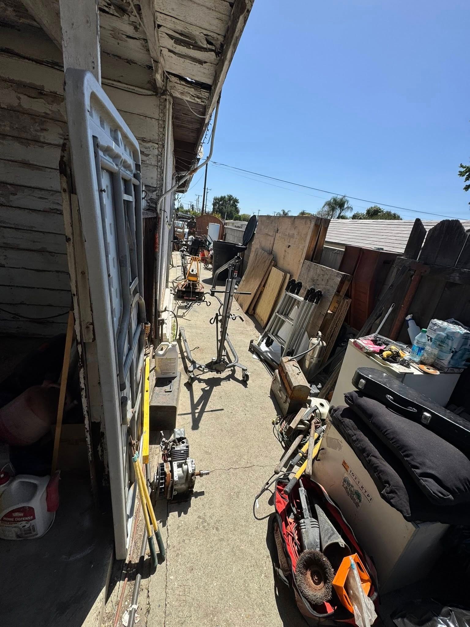 Narrow pathway cluttered with debris and building materials, flanked by structures. Sunny day.