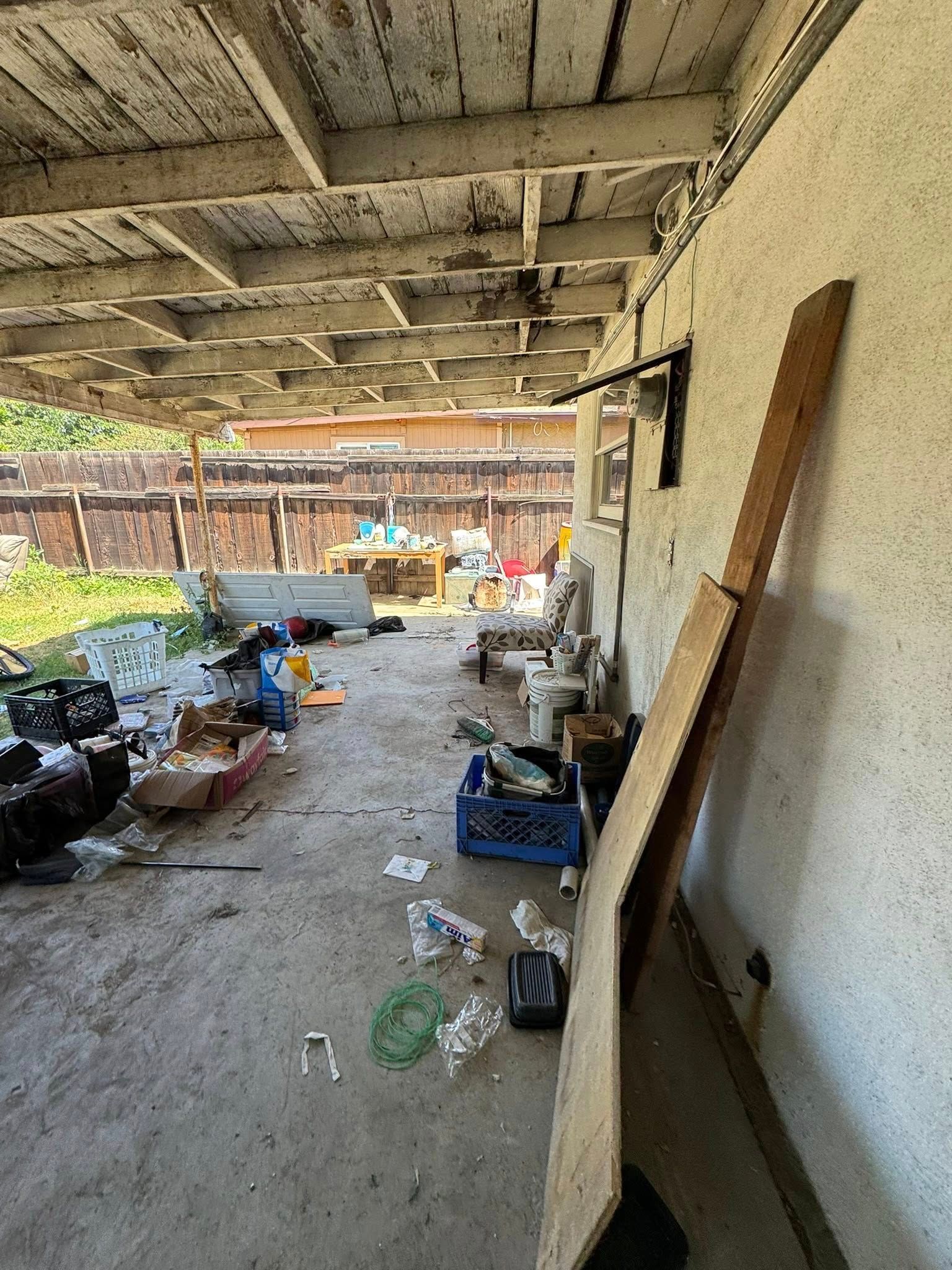 Cluttered outdoor patio under a deteriorating wooden roof. Trash and debris scattered on concrete floor.