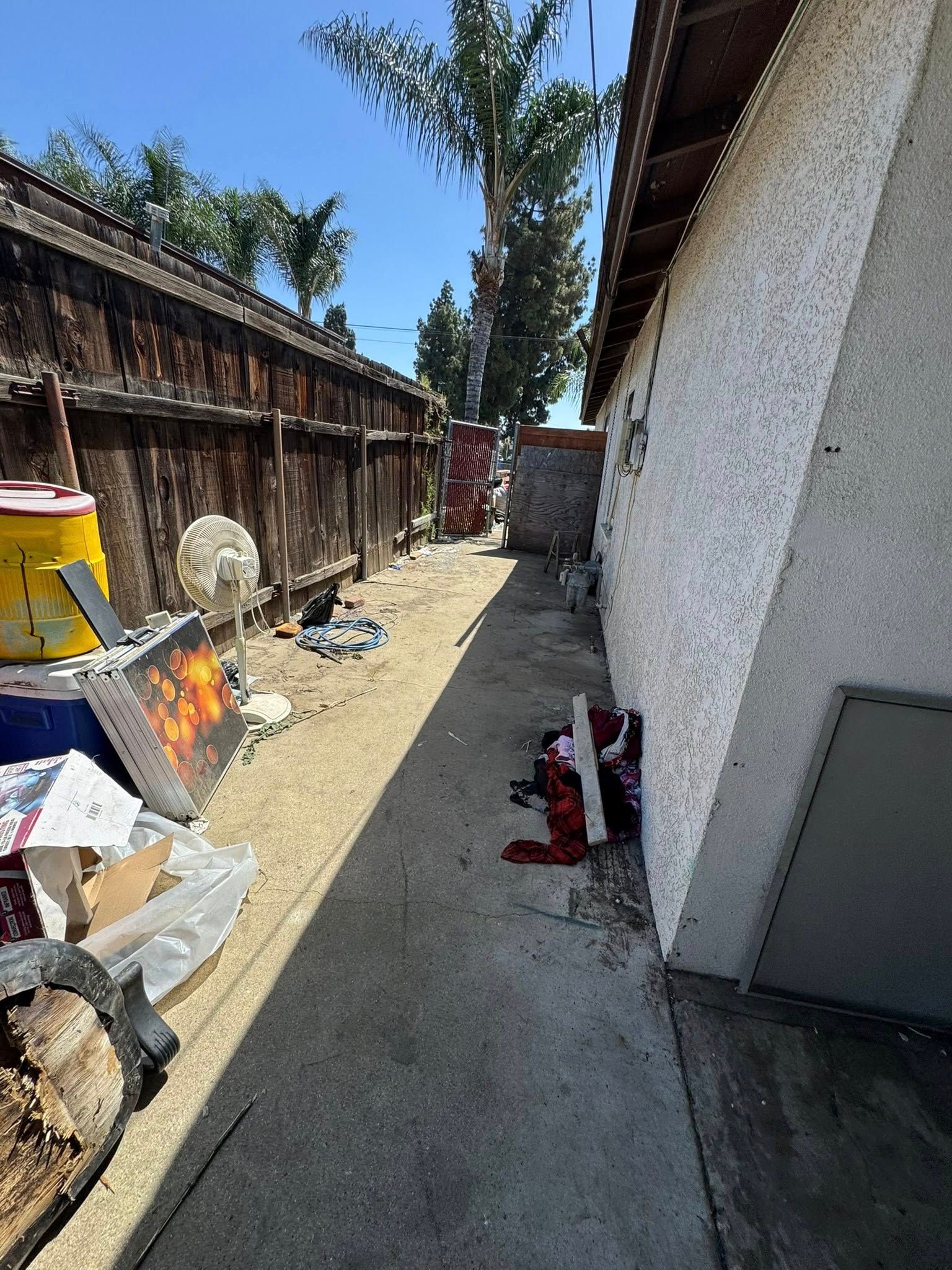 Narrow concrete walkway cluttered with trash and debris beside a wooden fence and building.