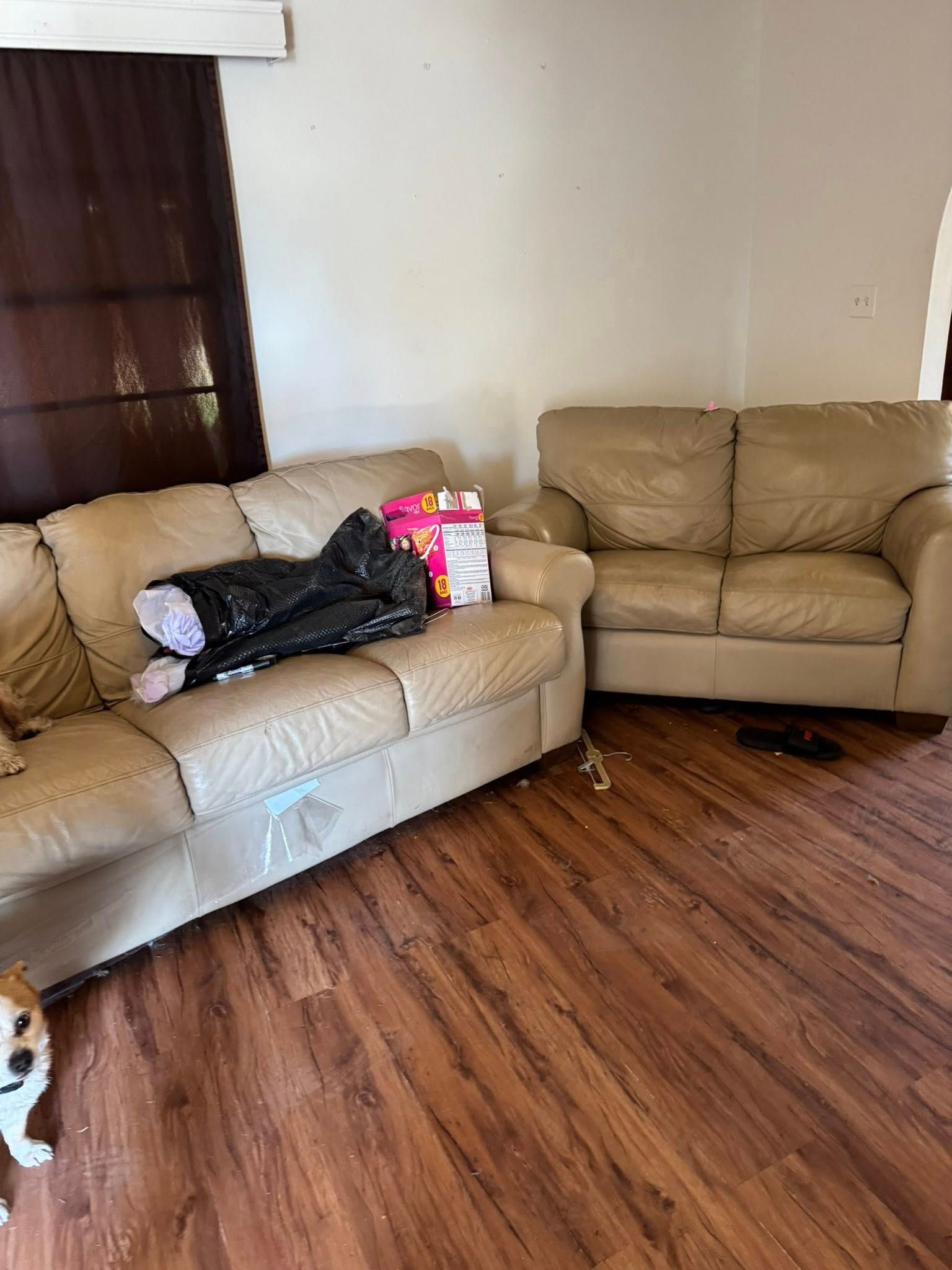 Two tan leather couches in a room with wood-look flooring and a window. Black bag and items on one couch.