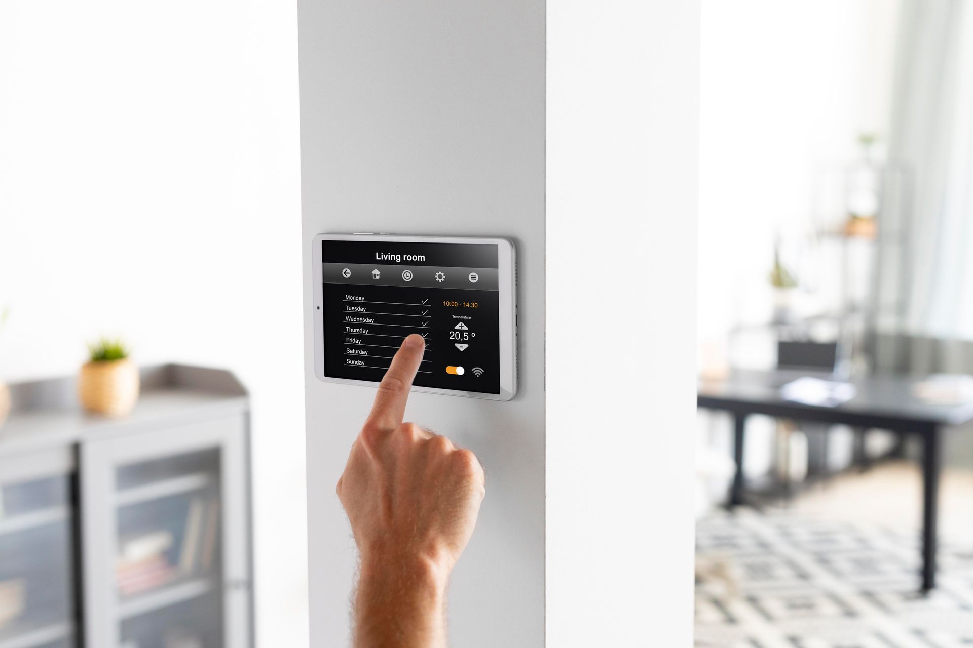 Hand tapping a wall-mounted touchscreen thermostat in a bright modern room