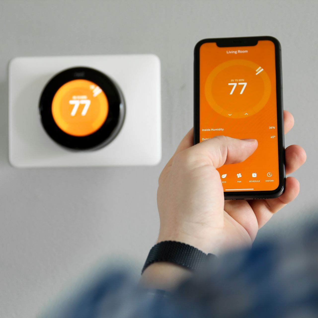 Person adjusting a smart thermostat on a wall via a smartphone. Both devices display the temperature, which is 77 degrees Fahrenheit.