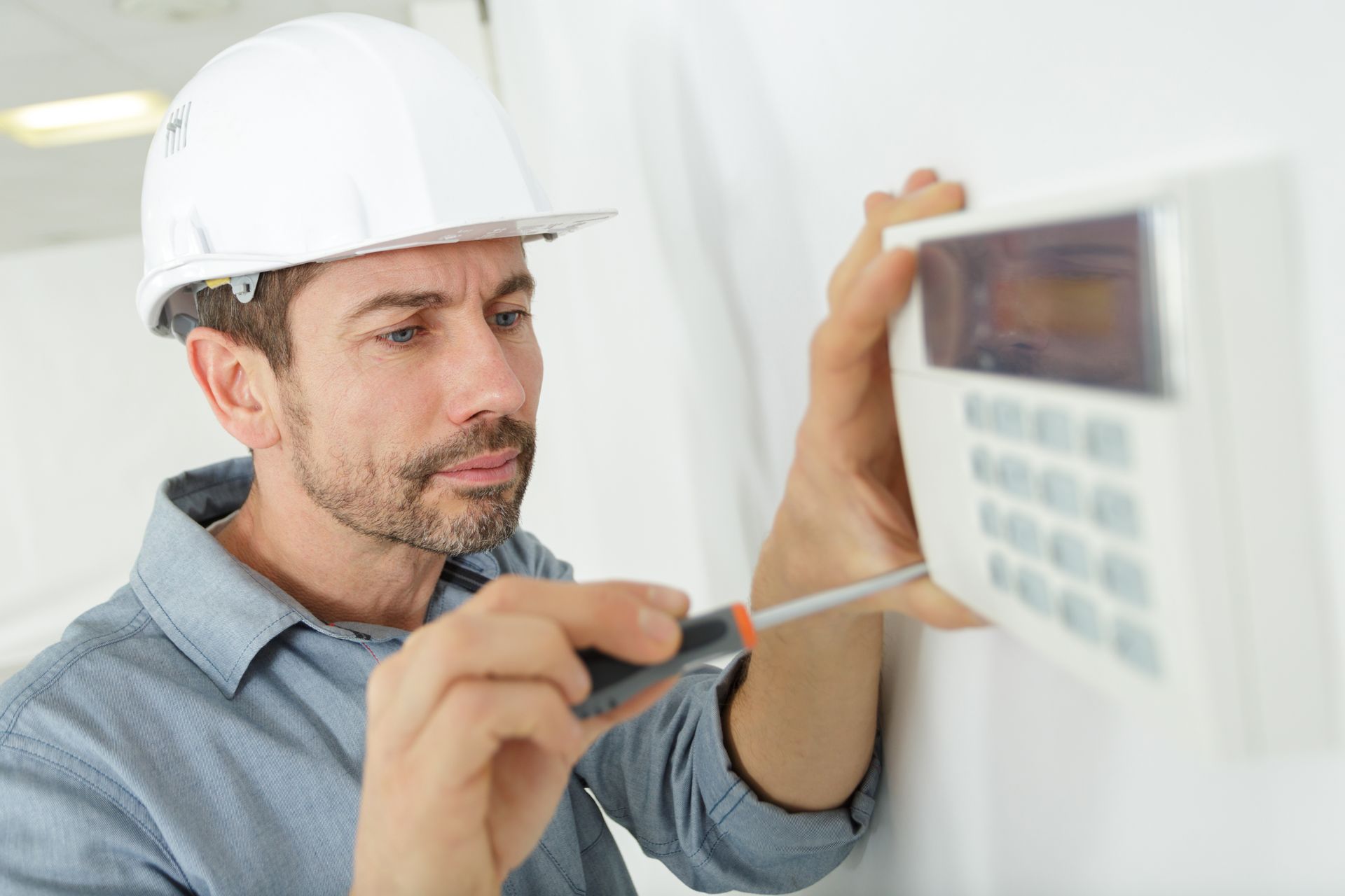 A man in a hard hat uses a screwdriver on a white security keypad on a wall. He appears focused.