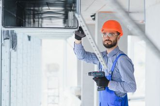 Construction worker in orange hard hat and blue overalls installs ductwork with a power drill.