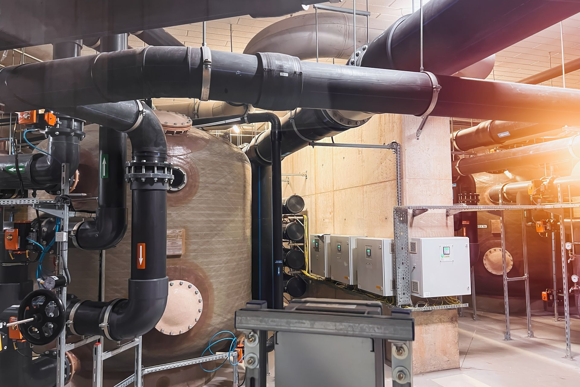 Industrial plant room with large black pipes, valves, and equipment under warm overhead lighting