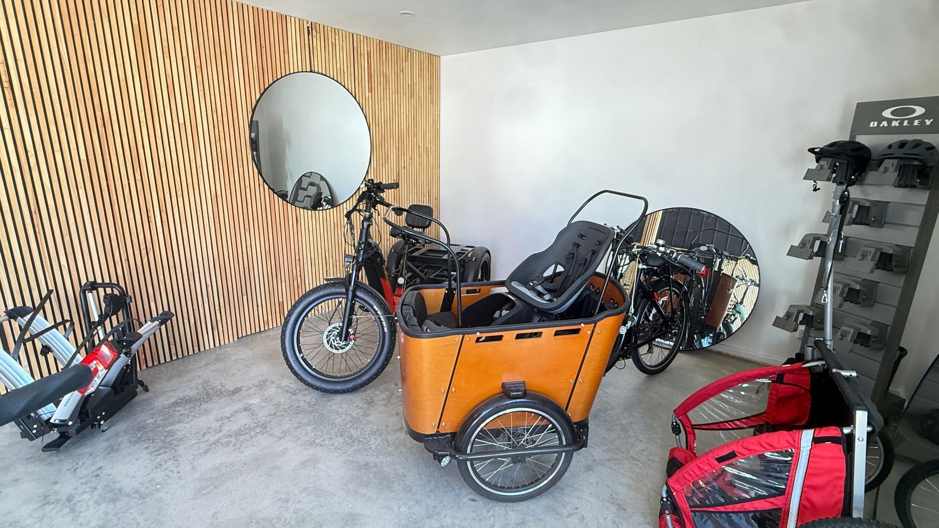 A garage space contains a cargo bike, two bicycles, a trailer, and a vertical display rack with bike helmets.