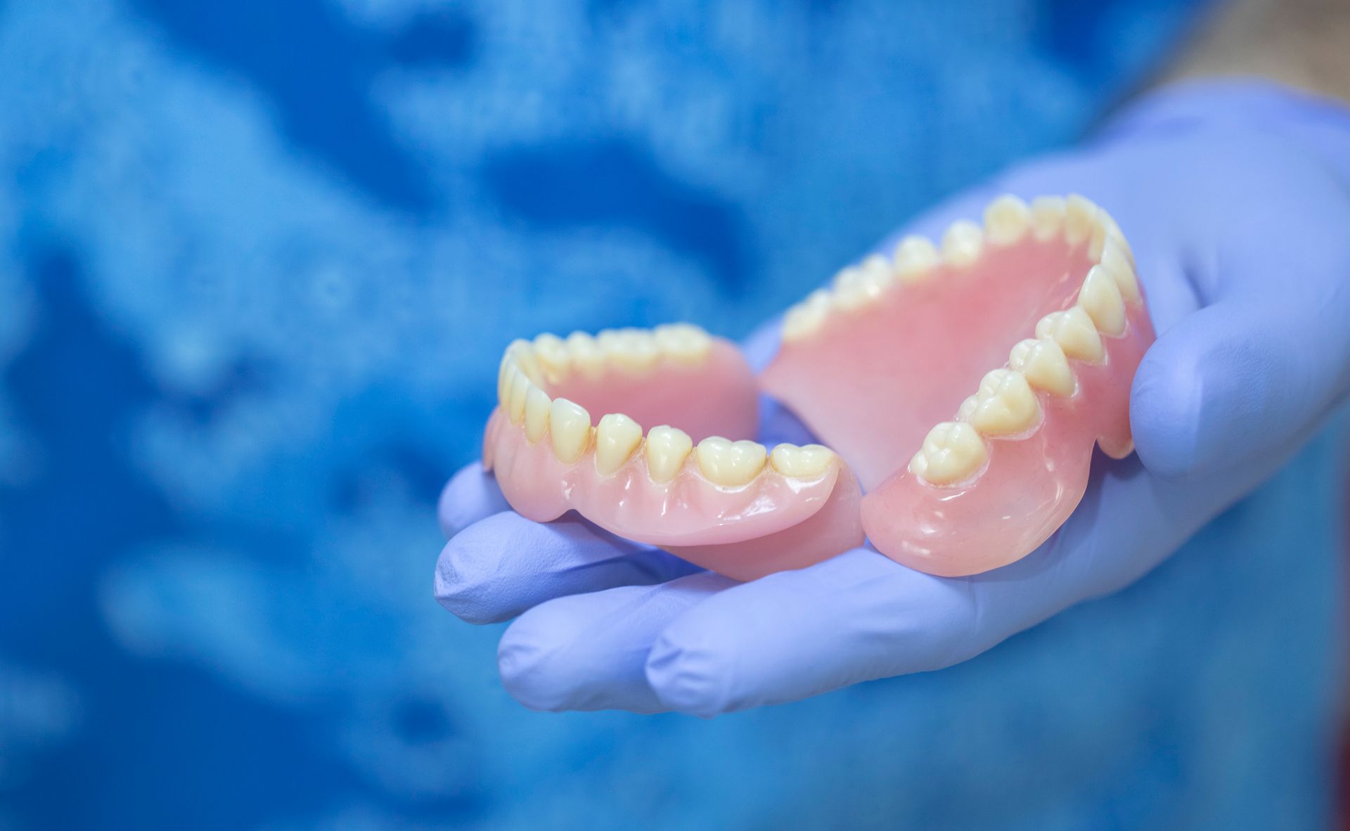 Dentures held in gloved hand, pink and yellow, against a blue background.