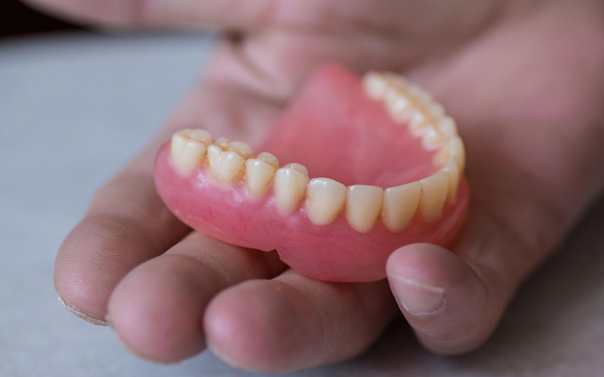 Hand holding a set of upper dentures; pink base with artificial teeth.