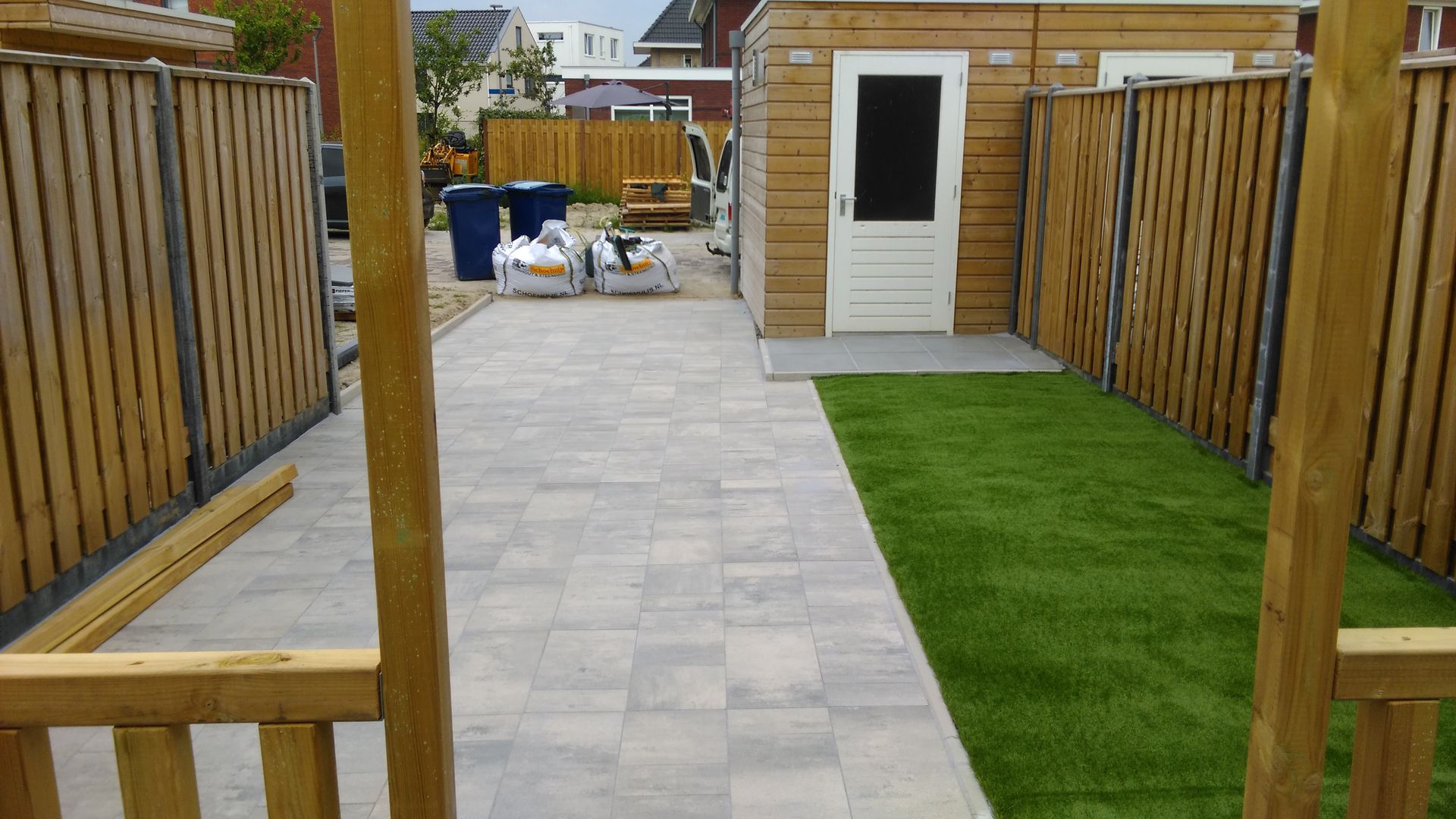 Terras in de achtertuin met grijze bestrating, groene gazonstroken, houten schuttingen en een klein schuurtje aan het einde.
