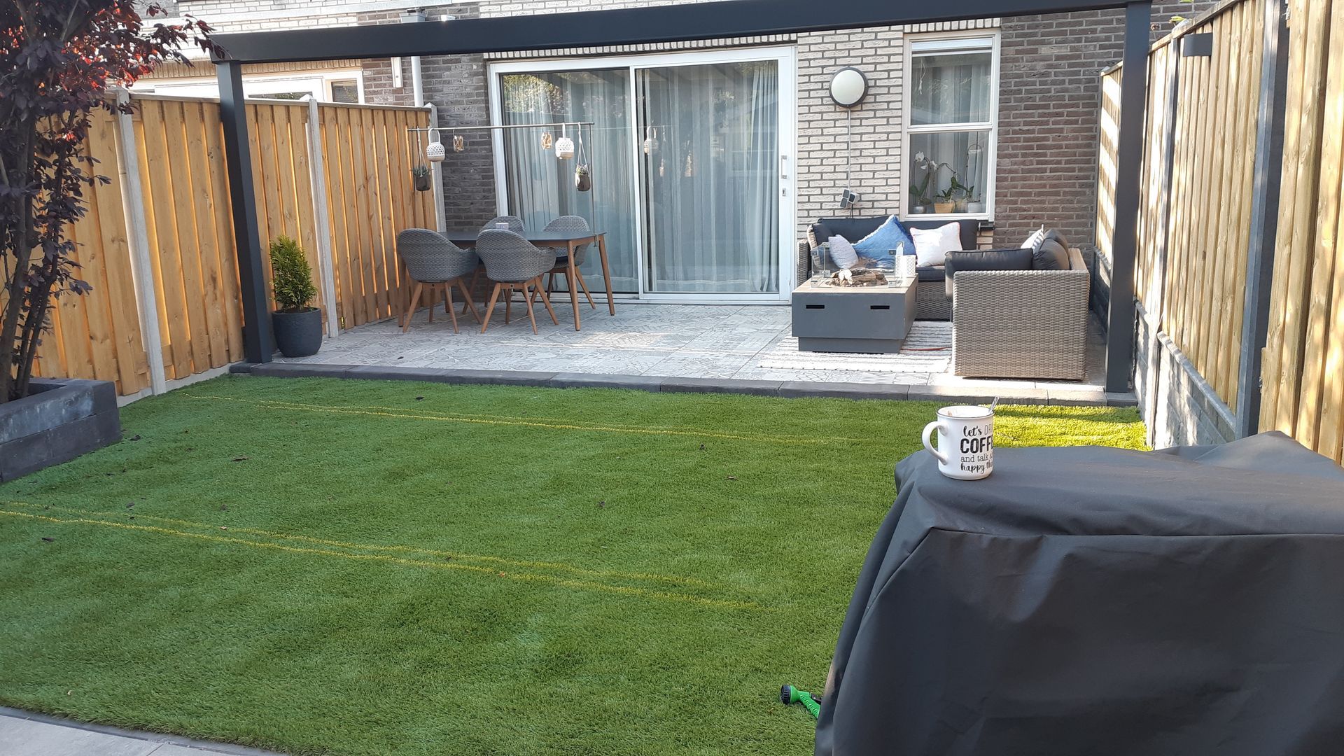 Terras in de achtertuin met gazon, rieten zitmeubelen en glazen schuifdeuren achter een houten schutting.