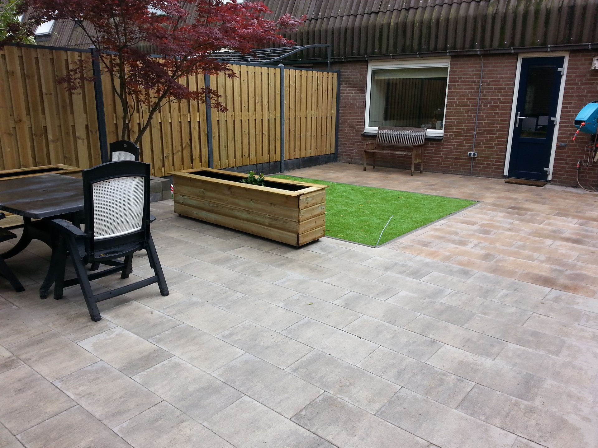 Terras met bakstenen huisje, betegeld zitgedeelte, verhoogde plantenbak en een klein gazon onder een boom met rode bladeren.