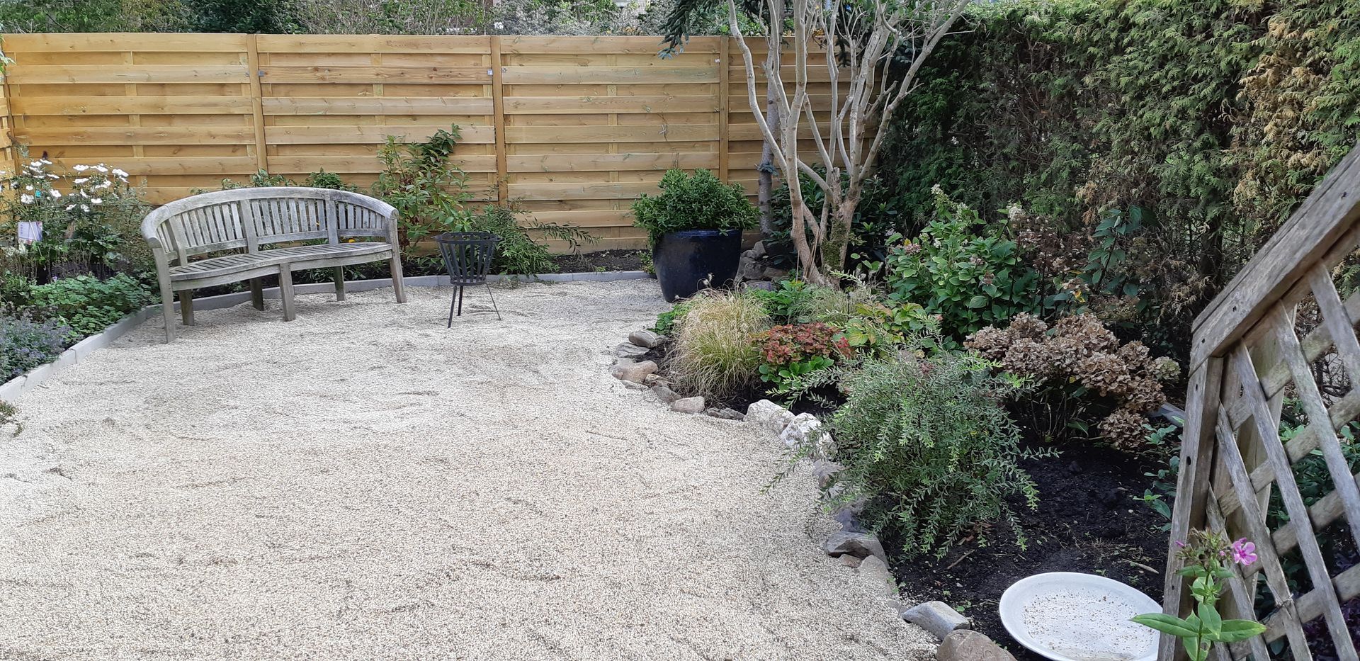 Een terras in de achtertuin met grind, een houten bank, een houten schutting en potplanten langs de rand.