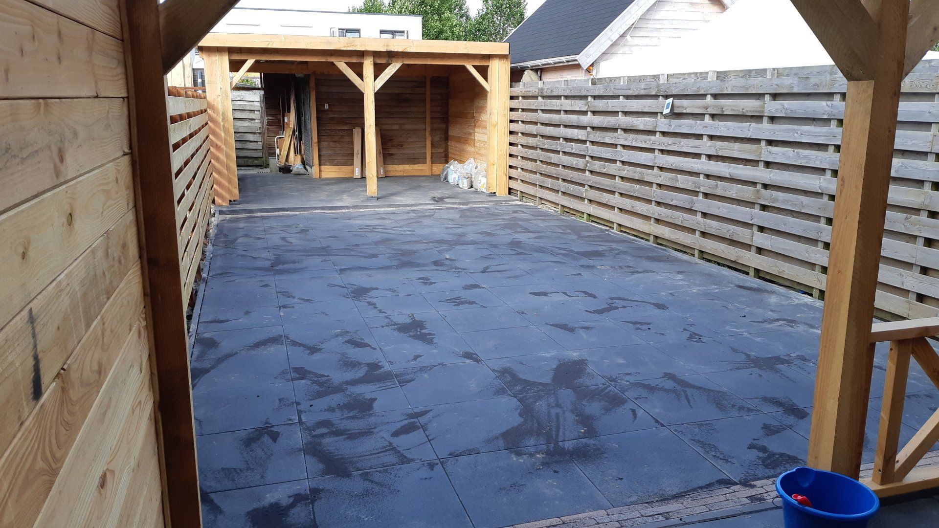 Overdekt terras in de achtertuin met een groot blauw zeildoek als vloer, houten schutting en een klein schuurtje aan het uiteinde.