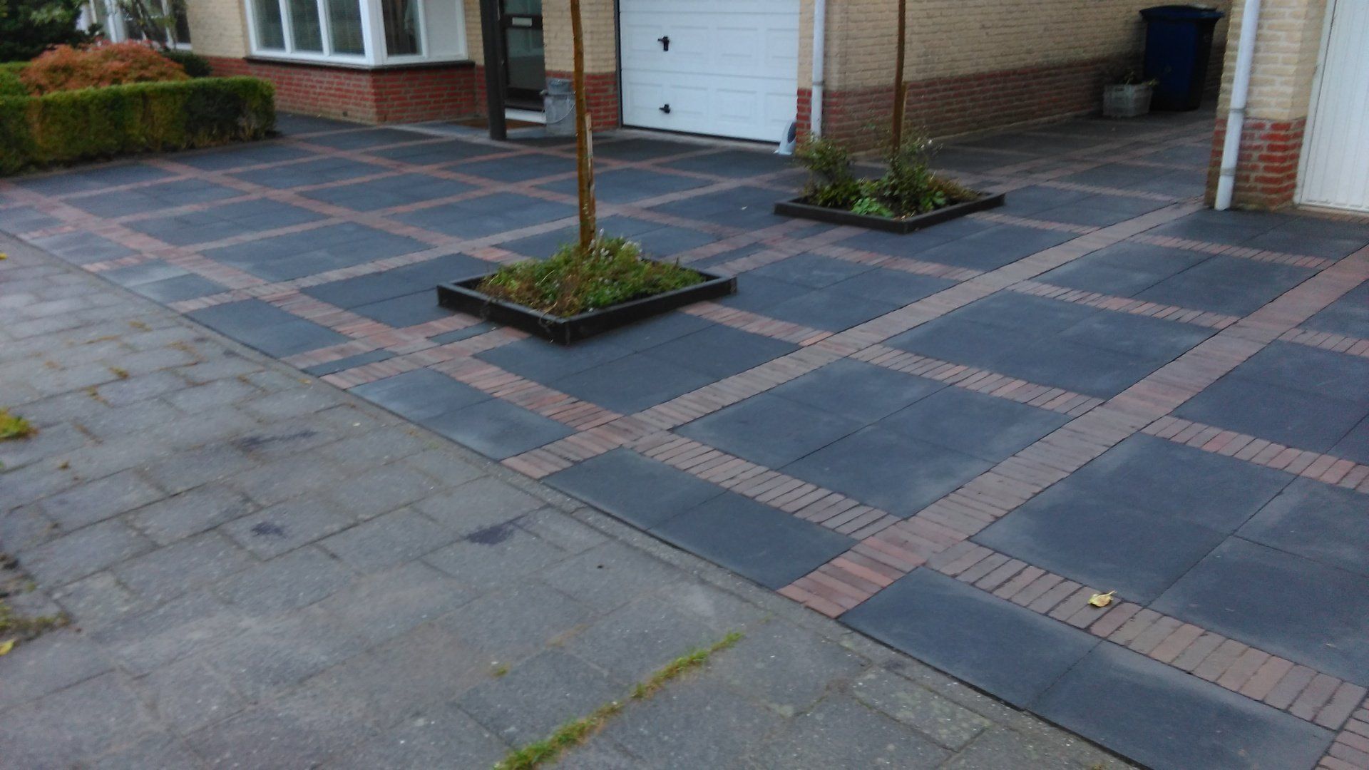 Een geplaveide oprit met een patroon van donkere en rode bakstenen leidt naar de ingang van het huis, met daaronder kleine aangeplante bomen.