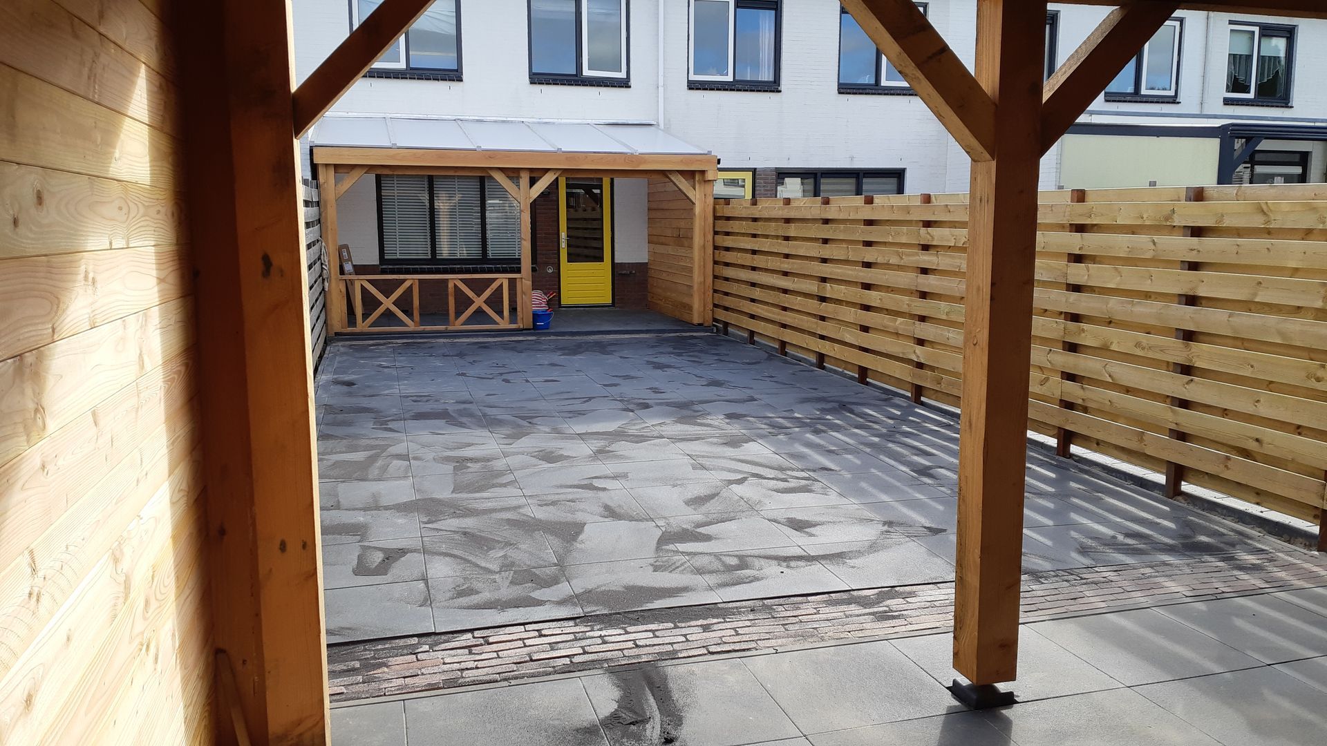 Overdekt terras in de achtertuin met bestrating, houten schutting en ramen onder een houten balkendak.