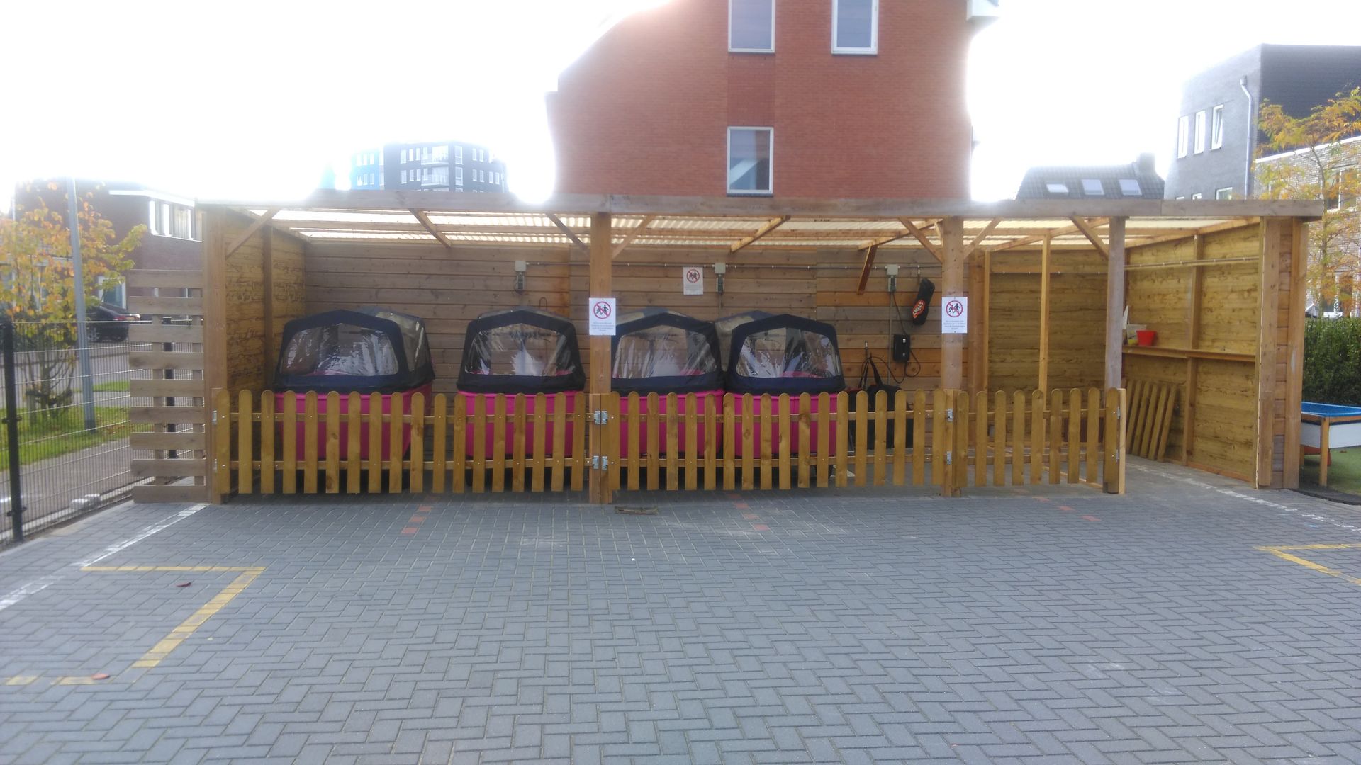 Overdekte fietsenstalling met meerdere fietsen achter een houten hek naast een bakstenen gebouw.