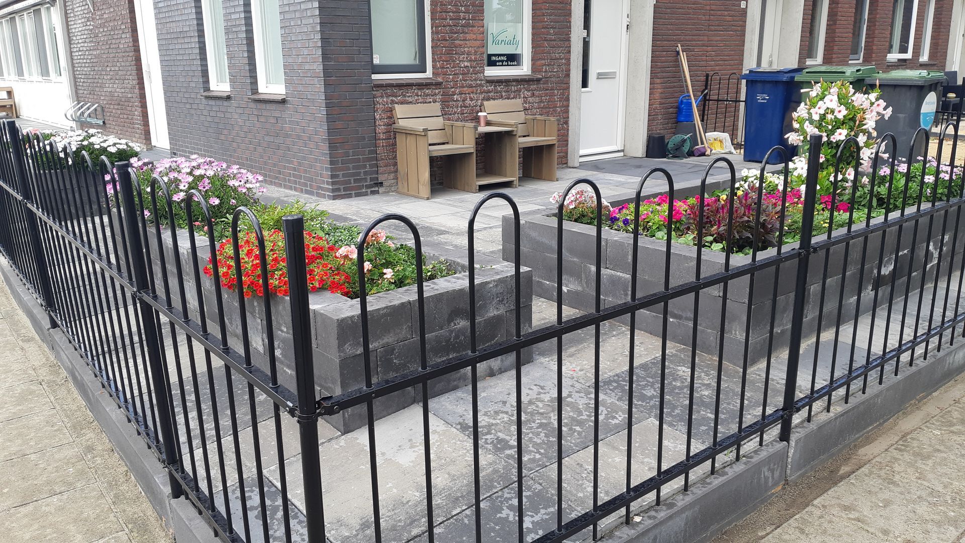 Een zwart metalen hek omringt een stenen plantenbak met kleurrijke bloemen naast een grijs bakstenen gebouw.