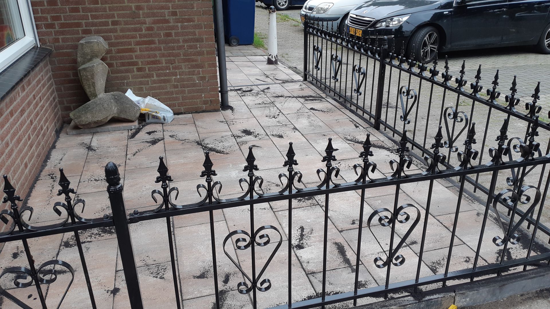 Een met sneeuw bedekte voortuin met een zwart ijzeren hek, een bakstenen muur en geparkeerde auto's op straat.