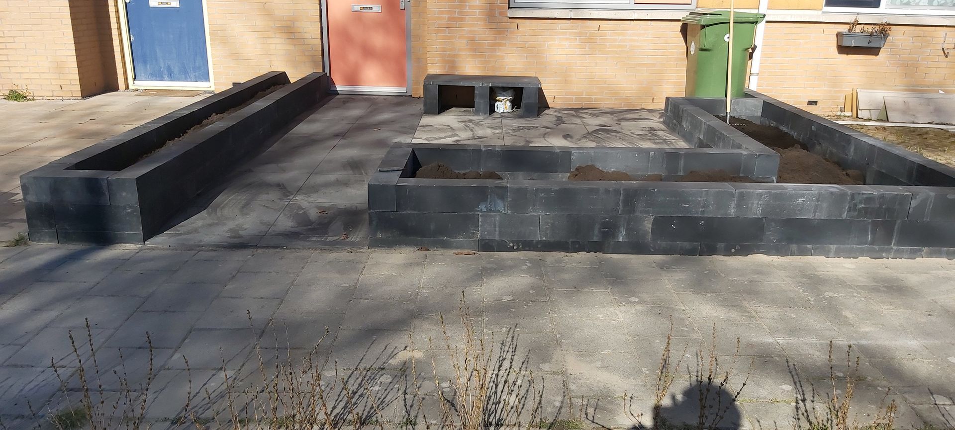 Een betonnen zitgedeelte buiten met zwarte, bankachtige wanden voor een gebouwingang.