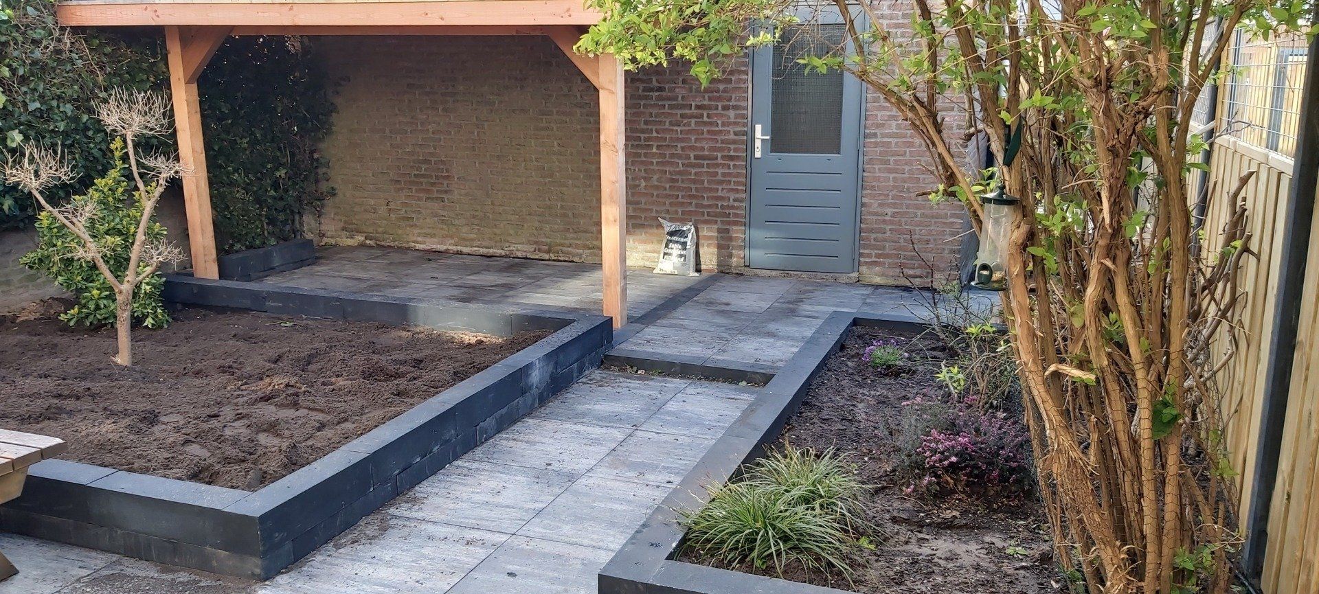 Tuinpad dat leidt naar een stenen veranda, met verhoogde bloembedden en struiken aan weerszijden.