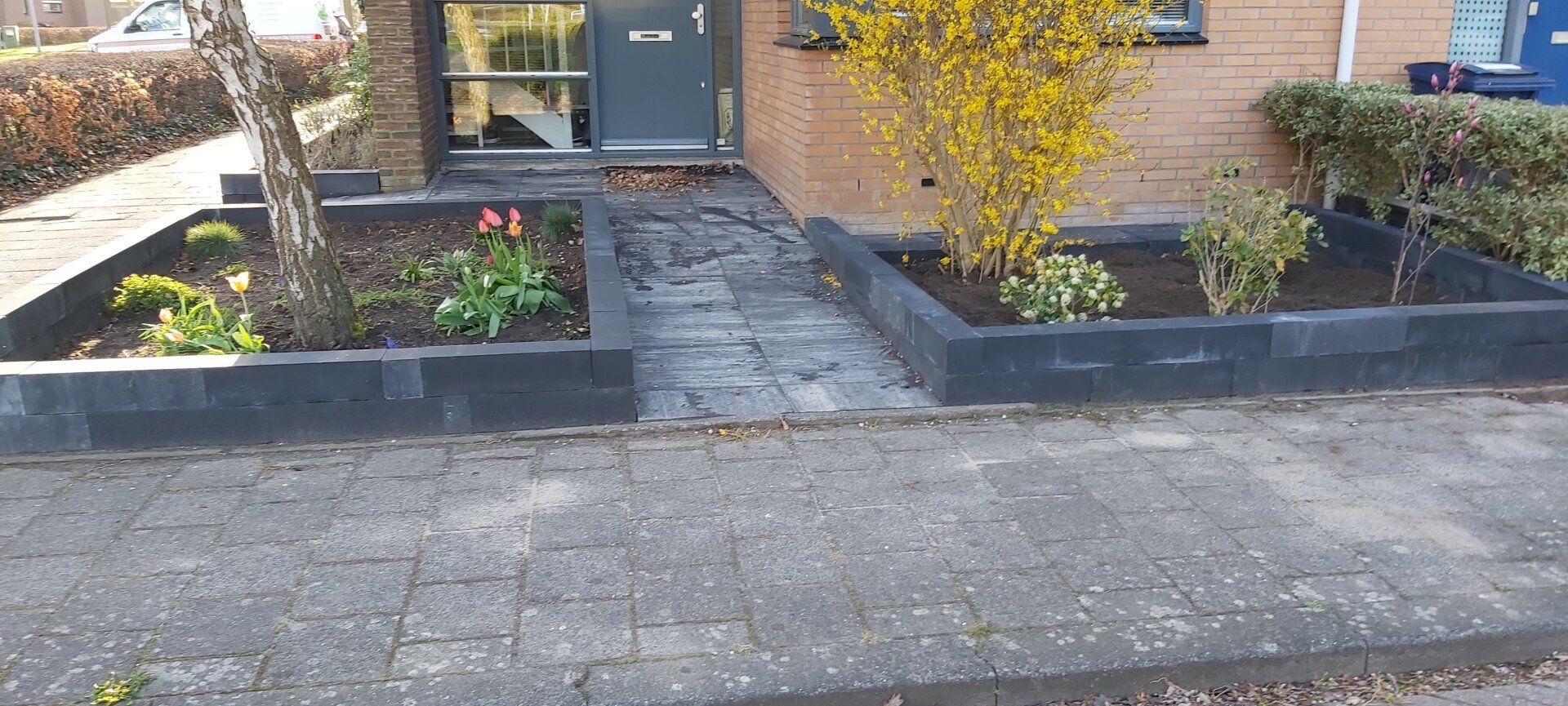 Voorpad met lage bakstenen plantenbakken dat naar de ingang van het huis leidt, begrensd door een stoeprand en oprit.