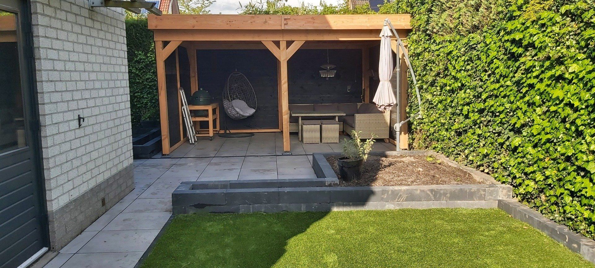 Terras in de achtertuin met houten overkapping, bakstenen muur, bestraat pad en groen gazon.