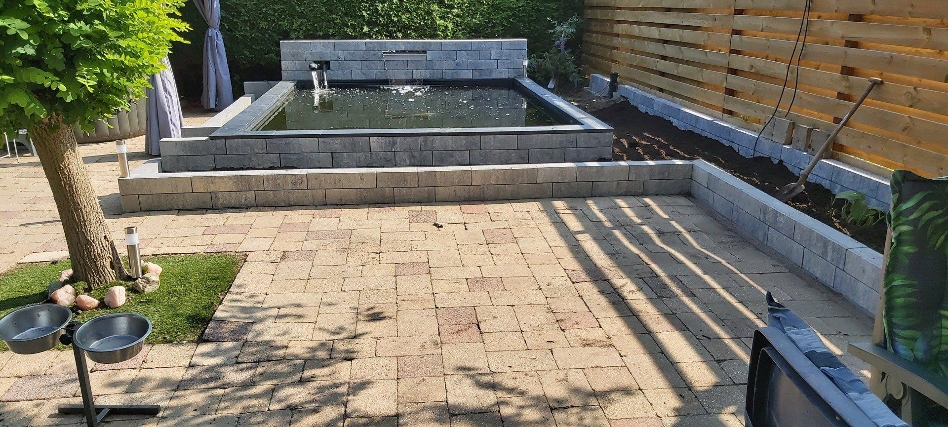 Terras in de achtertuin met stenen bestrating, verhoogd zwembad en houten schutting, badend in de zon.