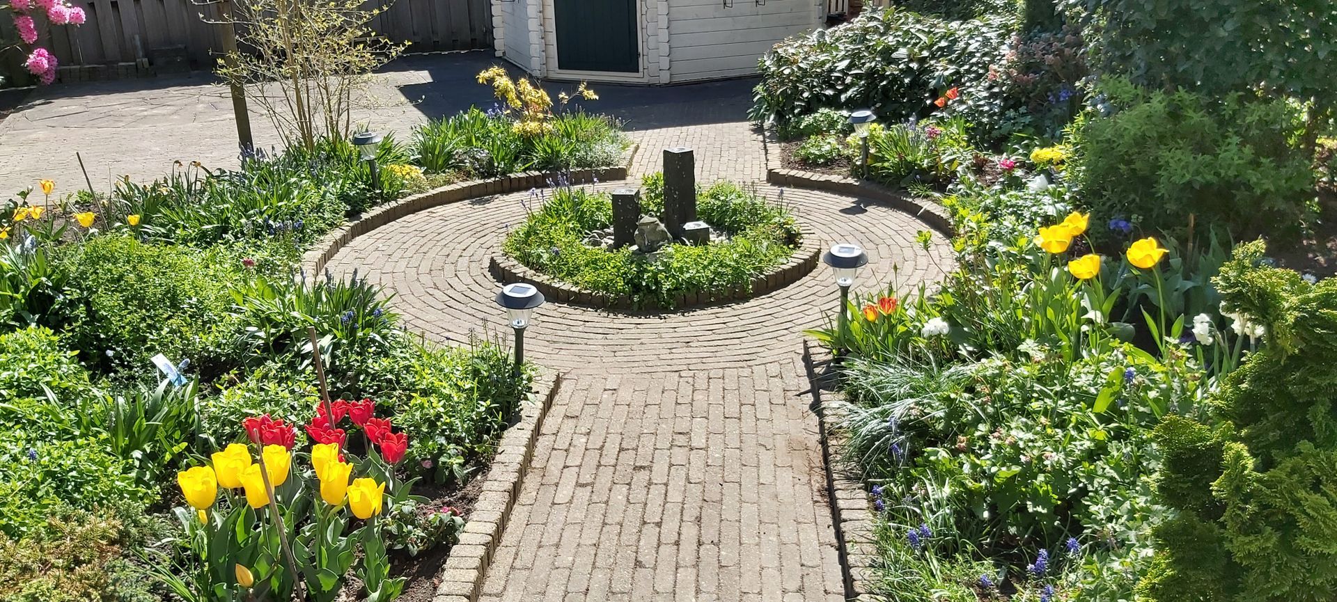 Een bakstenen pad door een kleurrijke bloementuin met een ronde stenen plantenbak in het midden.