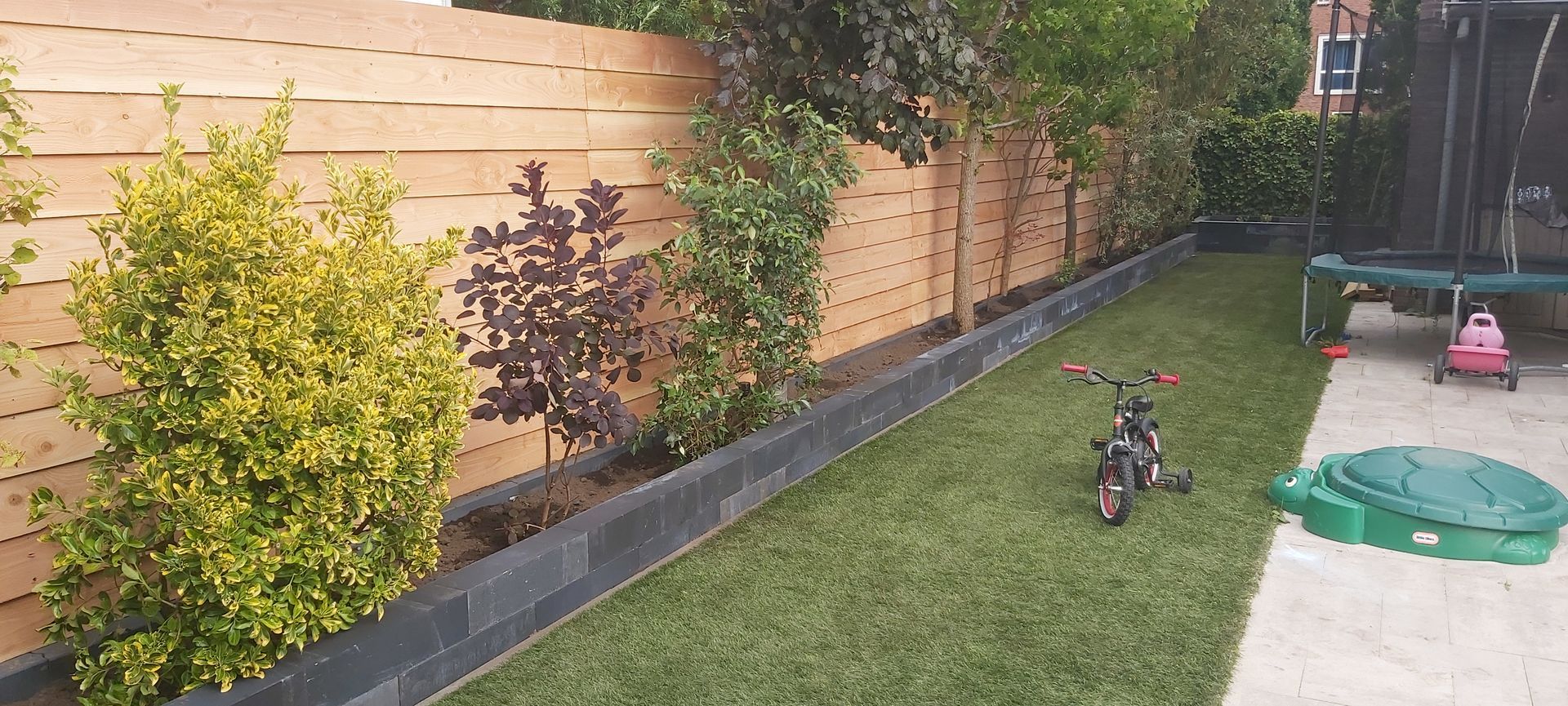 Gazon in de achtertuin naast een houten schutting met struiken, een kinderfietsje en een kleine speelset.