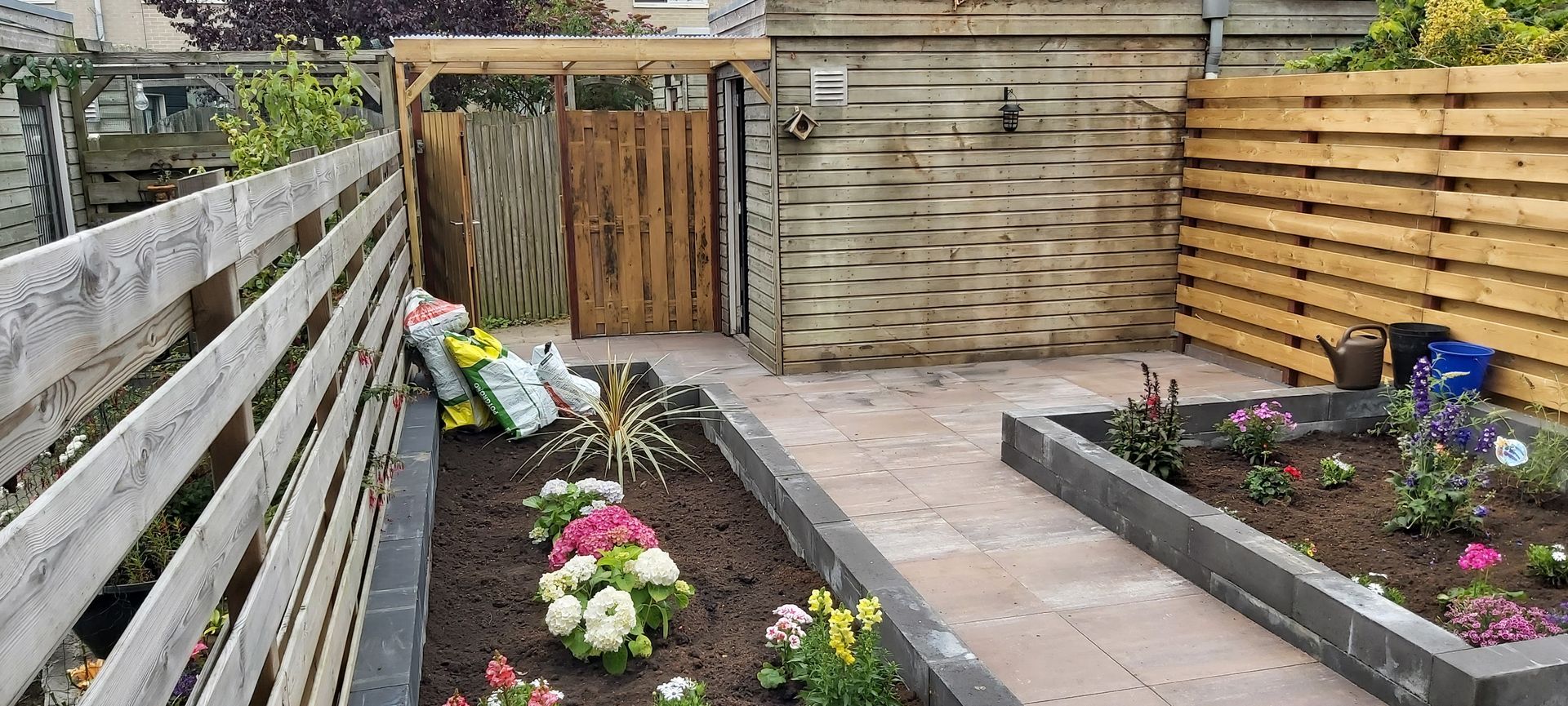 Achtertuin met verhoogde bloembedden, een betegeld terras, een houten schutting en een poort aan het einde.