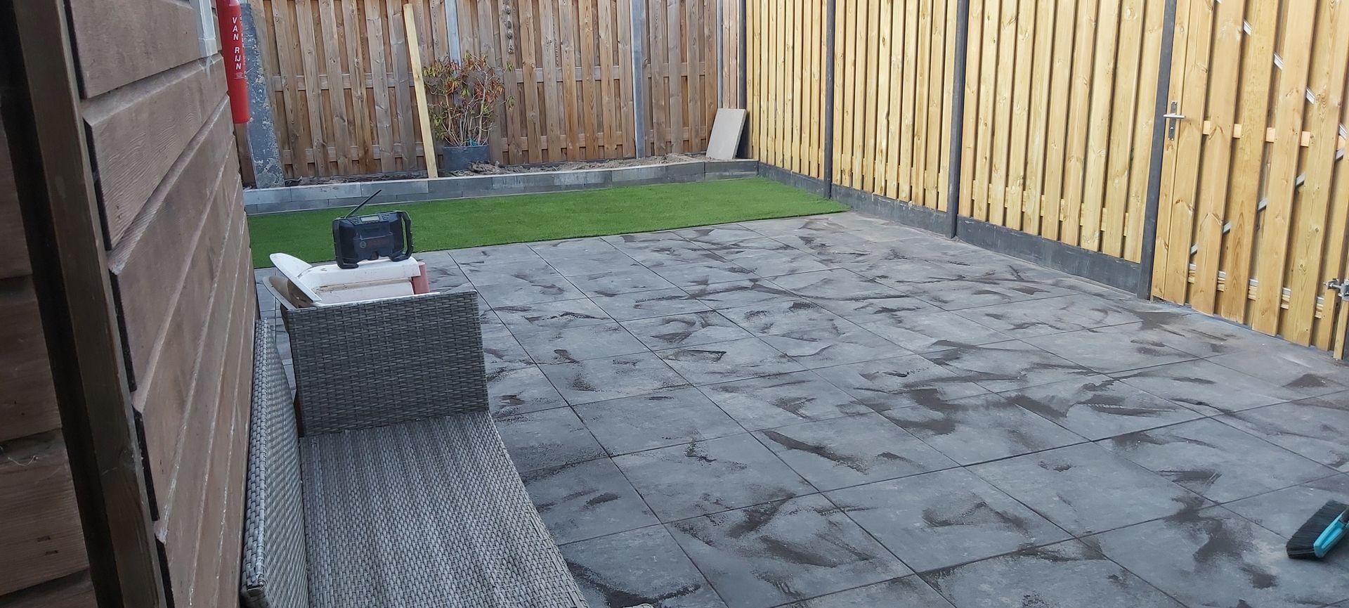 Klein, betegeld terras in de achtertuin met rieten zitmeubelen, een grasstrook en een houten schutting.