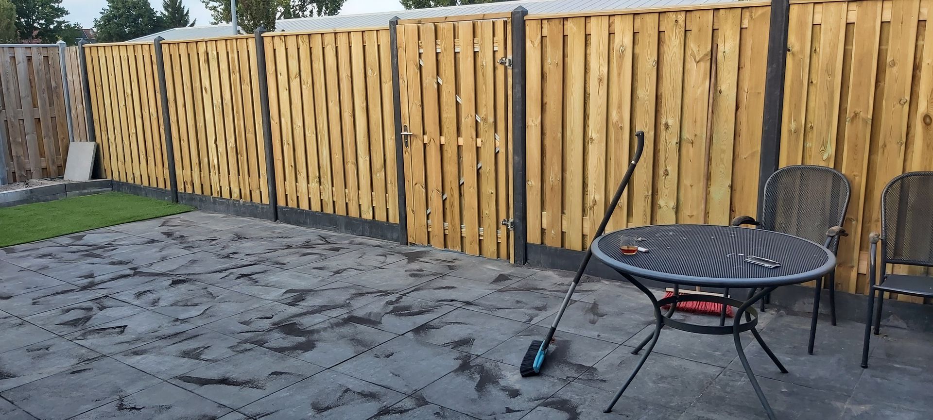 Terras in de achtertuin met een zwarte ronde tafel, twee stoelen en een houten schutting op een betonnen plaat.