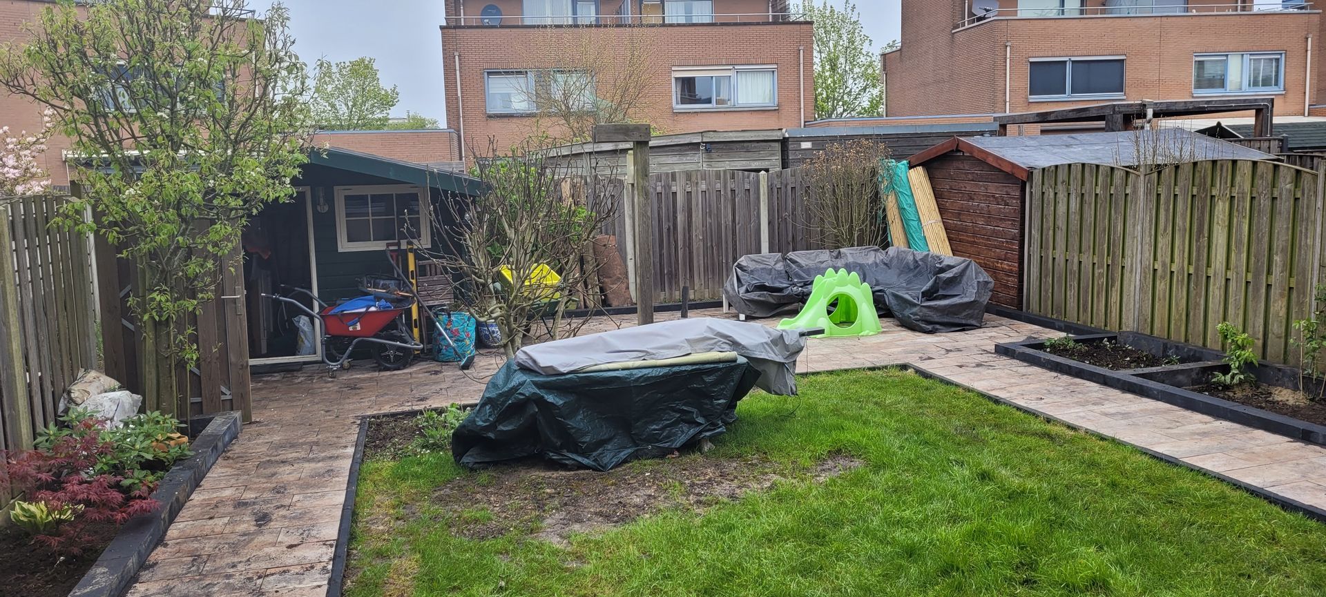 Achtertuin met overdekt terras, gazon, houten schutting en twee afgedekte spullen bij een schuur.