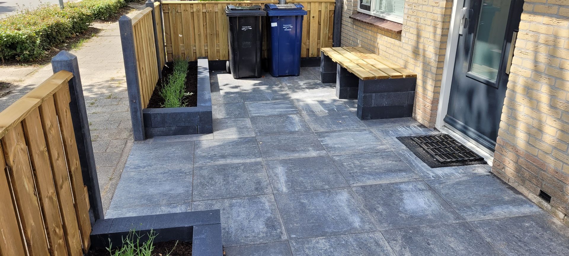 Terras in de achtertuin met grijze bestrating, houten schutting, bankjes en afvalbakken langs de achterwand.