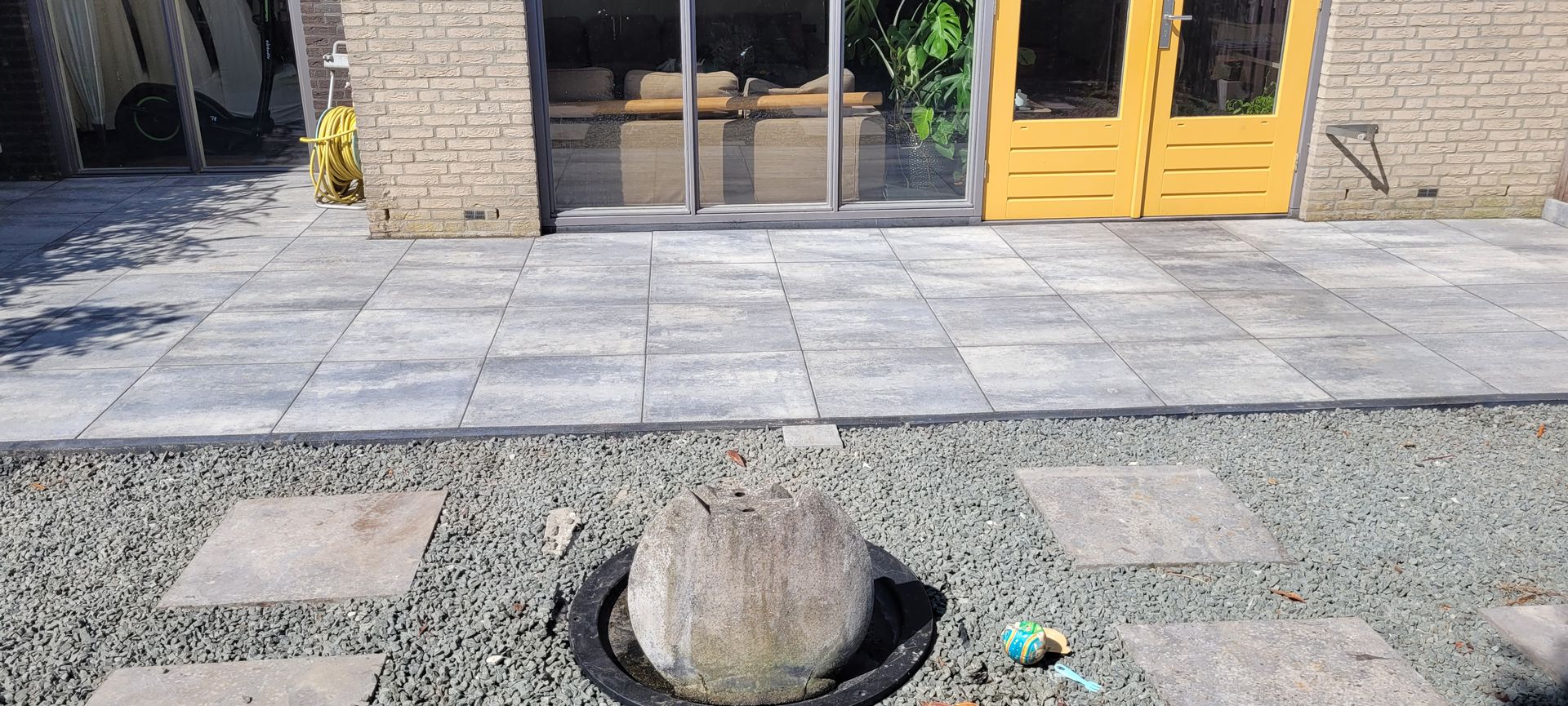 Een stoep met een boomstronk in een grindbed voor een gebouwingang met glazen deuren en gele kozijnen.