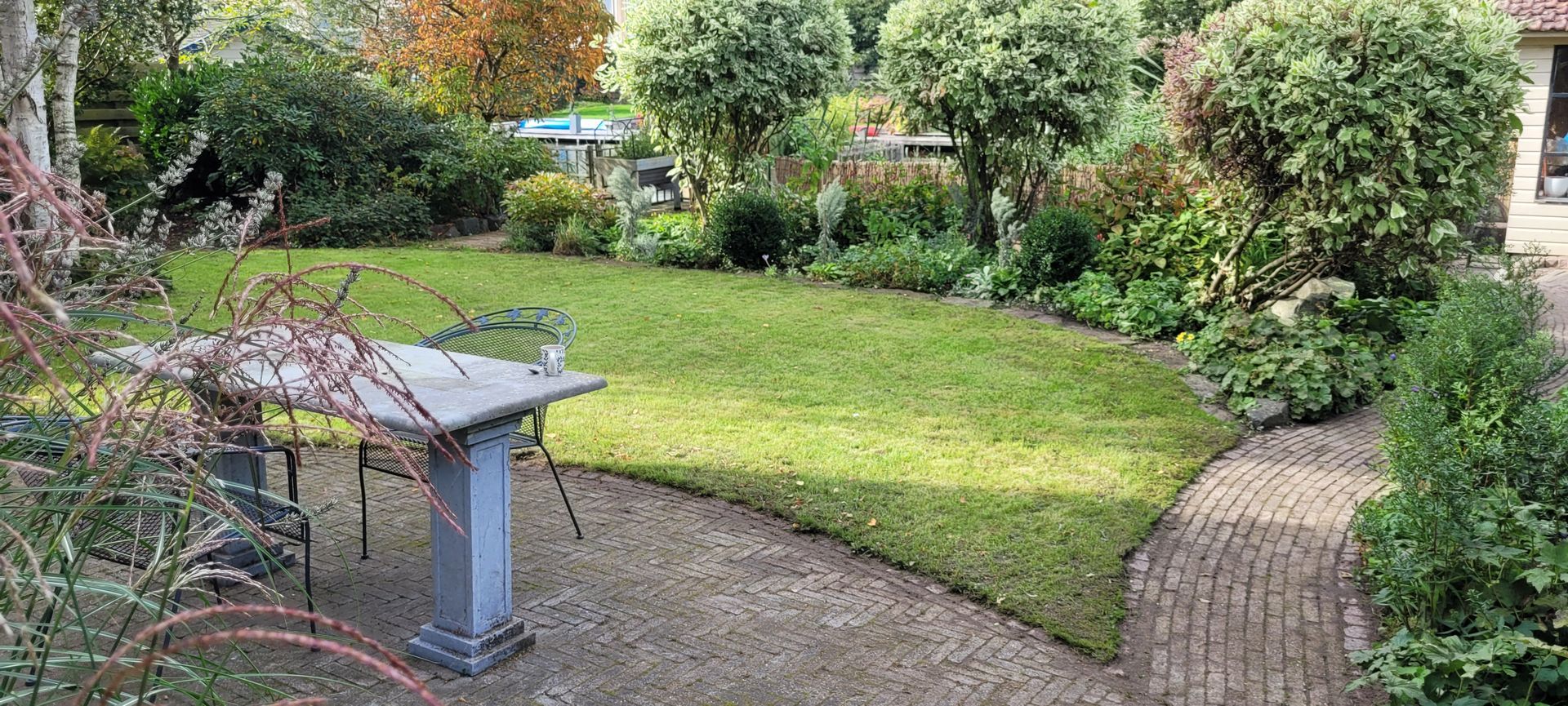 Achtertuin met gazon, struiken, bomen en een stenen tafel op een grindpad.