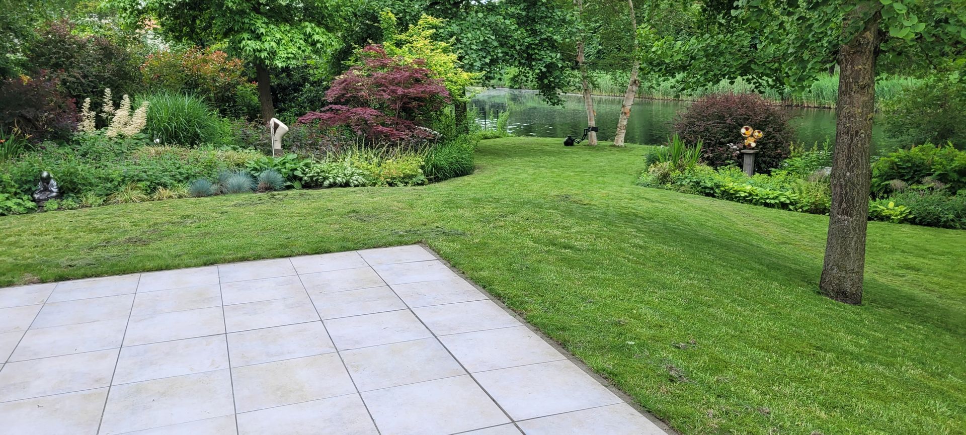 Tuinpatio met stenen tegels, groen gazon, struiken en bomen in een aangelegde tuin.