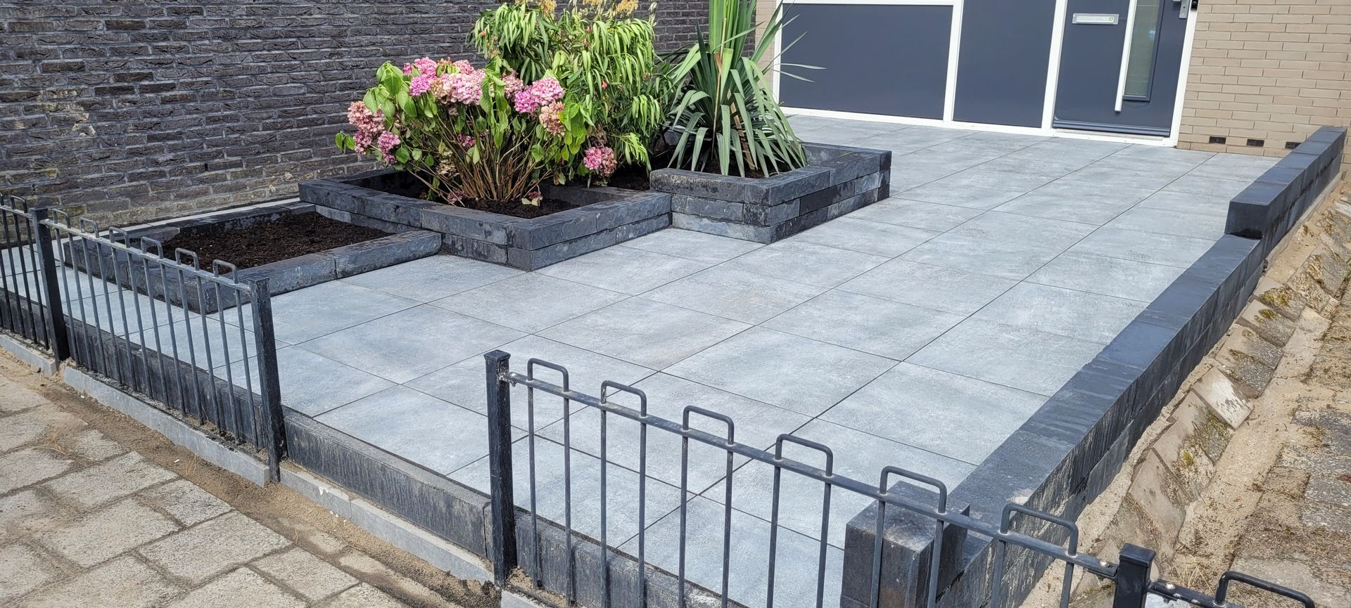 Verhoogd terras met grijze bestrating, zwarte metalen balustrade en een bloembed tegen een huismuur.