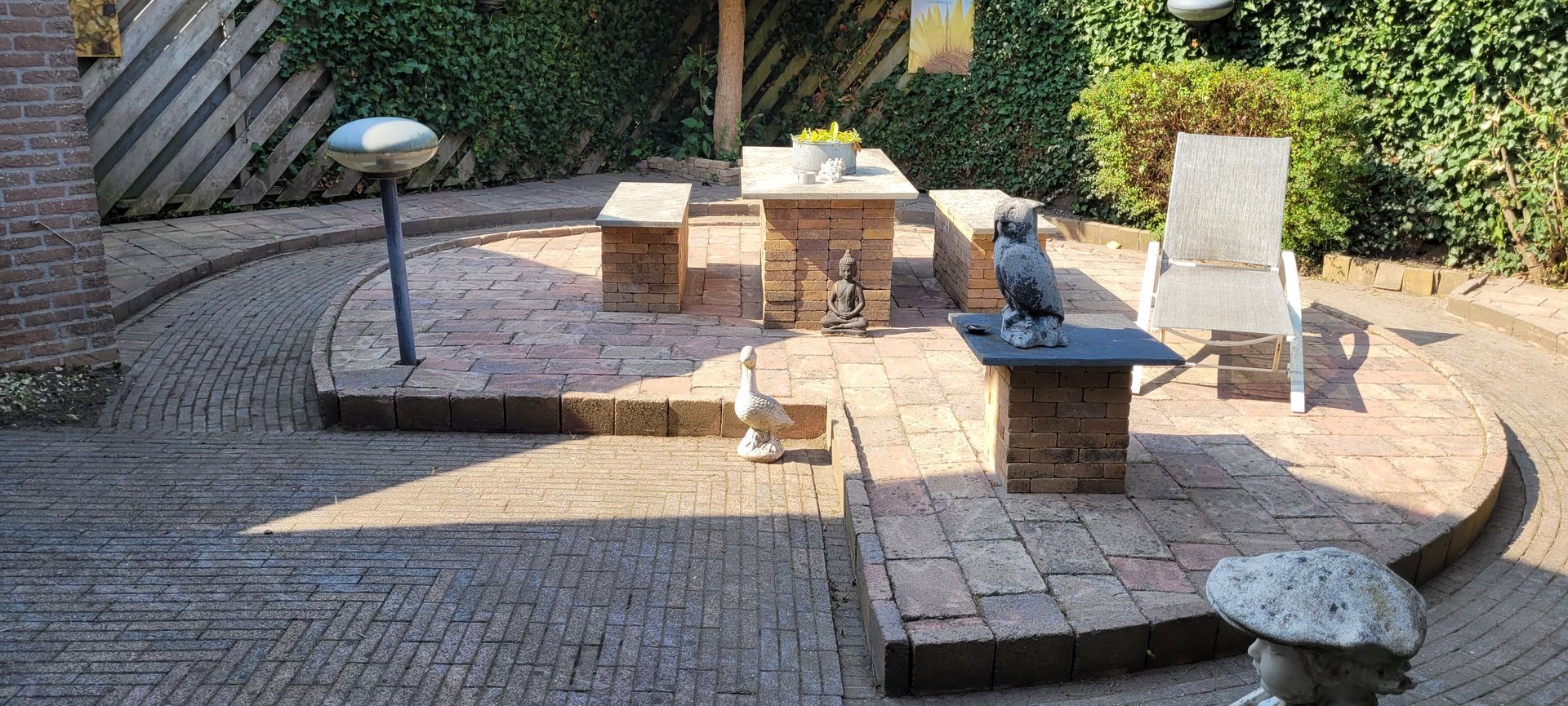 Stenen patio met bankjes, kleine tafels en een vogelbad in de zon.