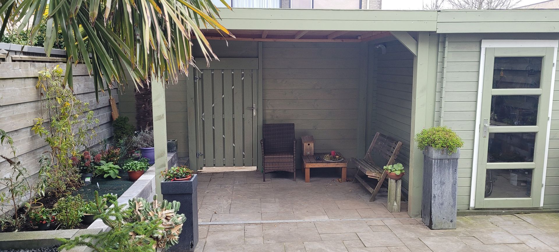 Overdekt terras met stoelen, potplanten en een tegelvloer naast een huis.