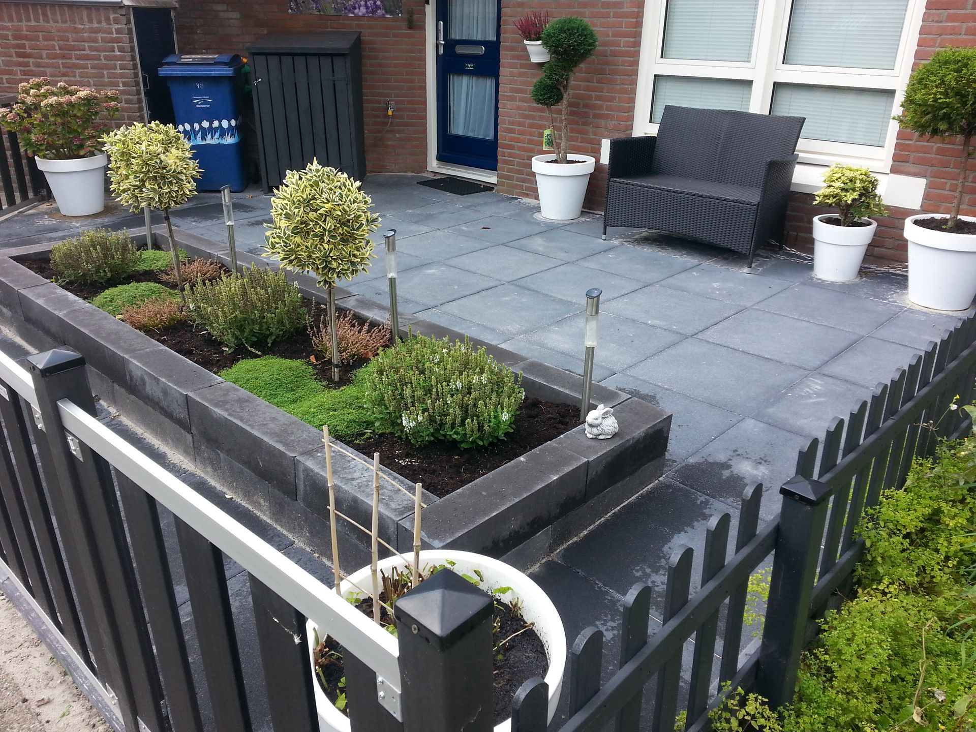 Klein terras in de voortuin met grijze bestrating, verhoogde plantenbakken, witte potten en een zwart hek bij de ingang van het huis.