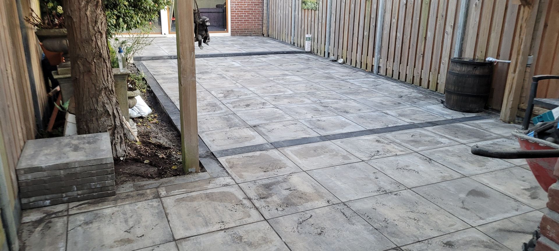 Betonnen terras met vierkante bestrating, houten schutting, boom en tuinafscheiding langs de zijkant van een tuin.