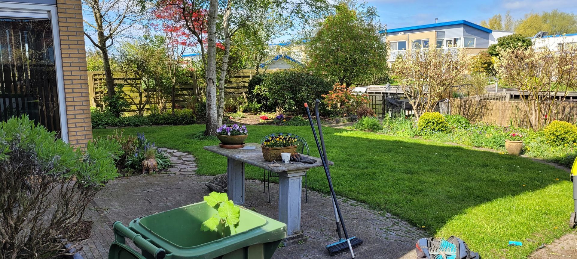 Zonnige achtertuin met terras, potplanten, gazon en een bankje bij een gebouw.