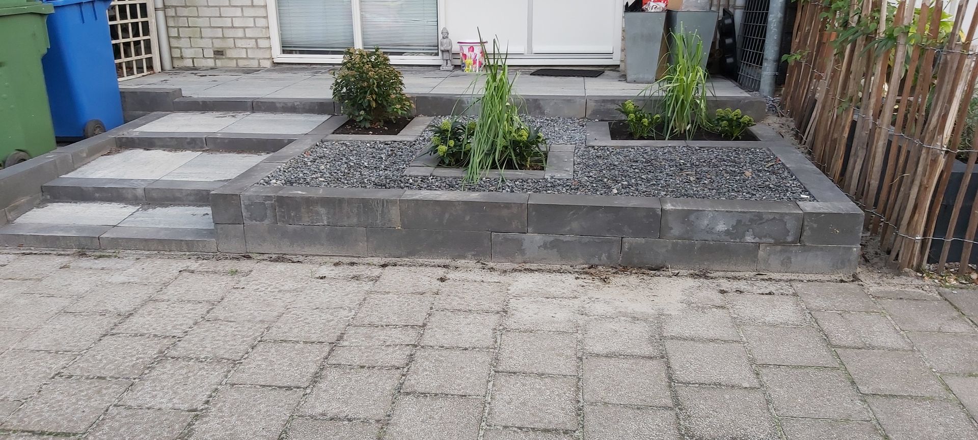 Kleine plantenbak in de voortuin met struiken en grind, naast een bestraat pad en de ingang van het huis.