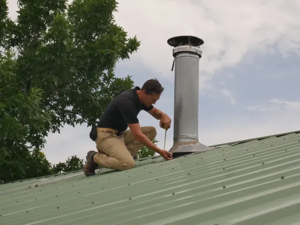 Worker kneels on a green metal roof, inspecting around a silver chimney.