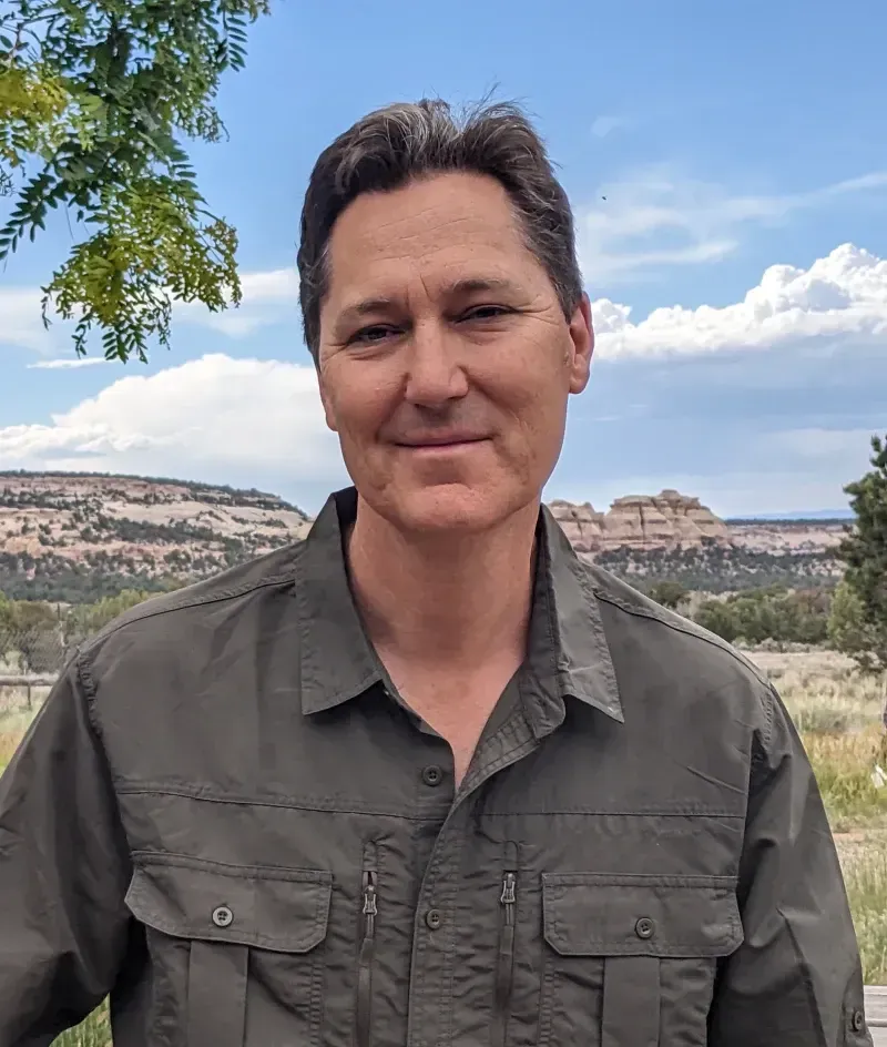Person standing outdoors in a green shirt, smiling with hills and blue sky in the background